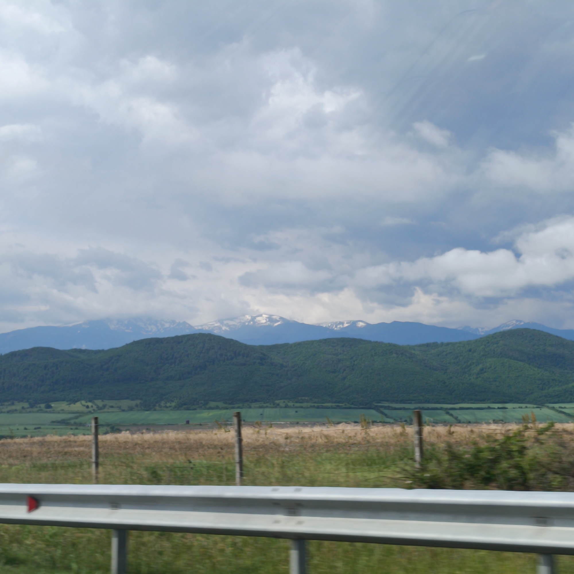 The image presents a broad landscape view, likely captured from a moving vehicle, featuring mountains, hills, fields, and an overcast sky. A silver metal guardrail, part of a roadside barrier, runs horizontally across the bottom section of the frame, with a small red triangular reflector visible on its left side.
Beyond the guardrail, the immediate foreground consists of dry, golden-brown grassy fields, intersected by a rustic fence with wooden posts and wire. Further into the mid-ground, there are expansive green agricultural fields and rolling hills densely covered in dark green forests.
In the distant background, majestic mountain ranges stretch across the horizon. Several of these peaks are distinguished by patches of white snow, indicating either high elevation or a cooler time of year. The sky above is largely covered with thick, gray and white clouds, creating diffused light, though hints of brighter sky can be seen in places. The overall impression is of a vast, natural, and somewhat rugged mountainous region under a cloudy daytime sky. The motion blur in the foreground suggests the photograph was taken while traveling. No visible text other than the red reflector.