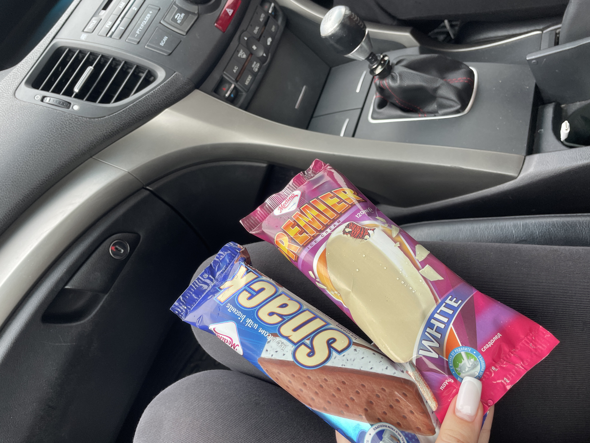The image presents a close-up view inside a car, likely in Dobromirtsi, Bulgaria, focusing on two ice cream bars held by a person's hand. The hand, adorned with long white fingernails, supports the two wrapped ice creams against a dark grey fabric background, likely a lap or seat.
The left ice cream bar is in a blue package, prominently displaying the brand "Snack" and describing its contents as "ice cream with biscuits." Additional text includes "Medina" and "REAL MILK FROM RHODOPES." The right ice cream bar is encased in a vibrant pink and purple wrapper. It is branded "PREMIER" and identified as "WHITE ICE CREAM," with a volume of "120ml e." The packaging also features "сладолед" (Bulgarian for ice cream) and "παγωτό" (Greek for ice cream), along with "С МЛЯКО ОТ РОДОПИТЕ" (with milk from the Rhodopes), indicating local sourcing from the Rhodope mountains.
The car's interior serves as the background, revealing a grey and black dashboard. On the left, an air conditioning vent with a metallic adjuster is visible, alongside various audio controls such as "TITLE," "FM," "AM," "SCAN," and "RETURN." To the right, a manual transmission gear shifter with a black knob and red-stitched leather boot is prominent. Below the shifter, climate control buttons, including "A/C," "DUAL AUTO," and "MODE," can be seen. The scene depicts a casual, daytime moment, possibly a break during travel, where cool refreshments are being enjoyed.