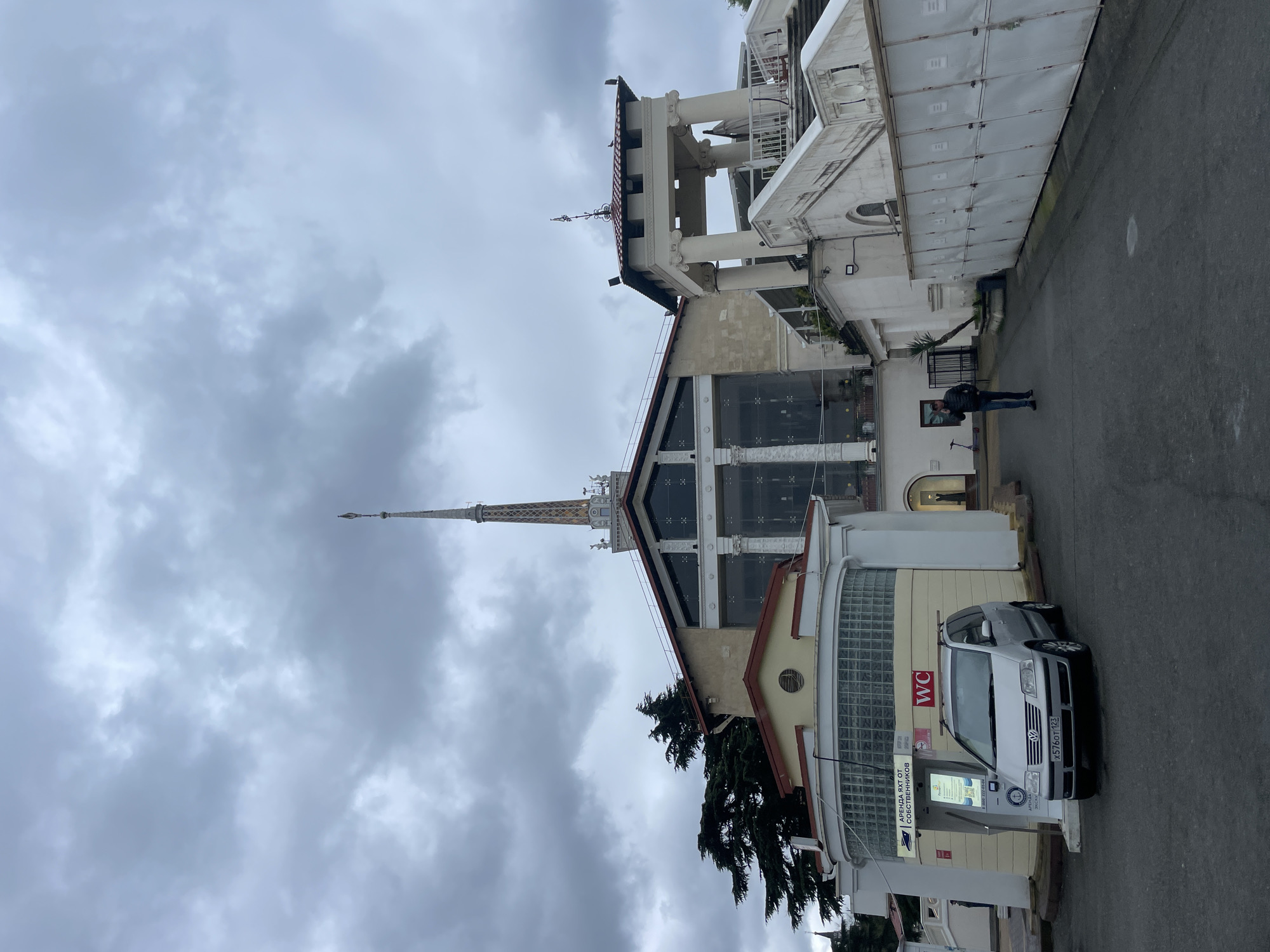 The image shows a street scene in Sochi, Russia. The sky is overcast with dense, grey clouds. On the right side of the frame is a multi-story building with a variety of architectural styles, including classical columns and modern glass paneling. A spire extends from the top of the building toward the sky. A man is walking on the street, away from the camera. A silver car is parked next to a building with a "WC" sign visible. A parking kiosk is next to the car.