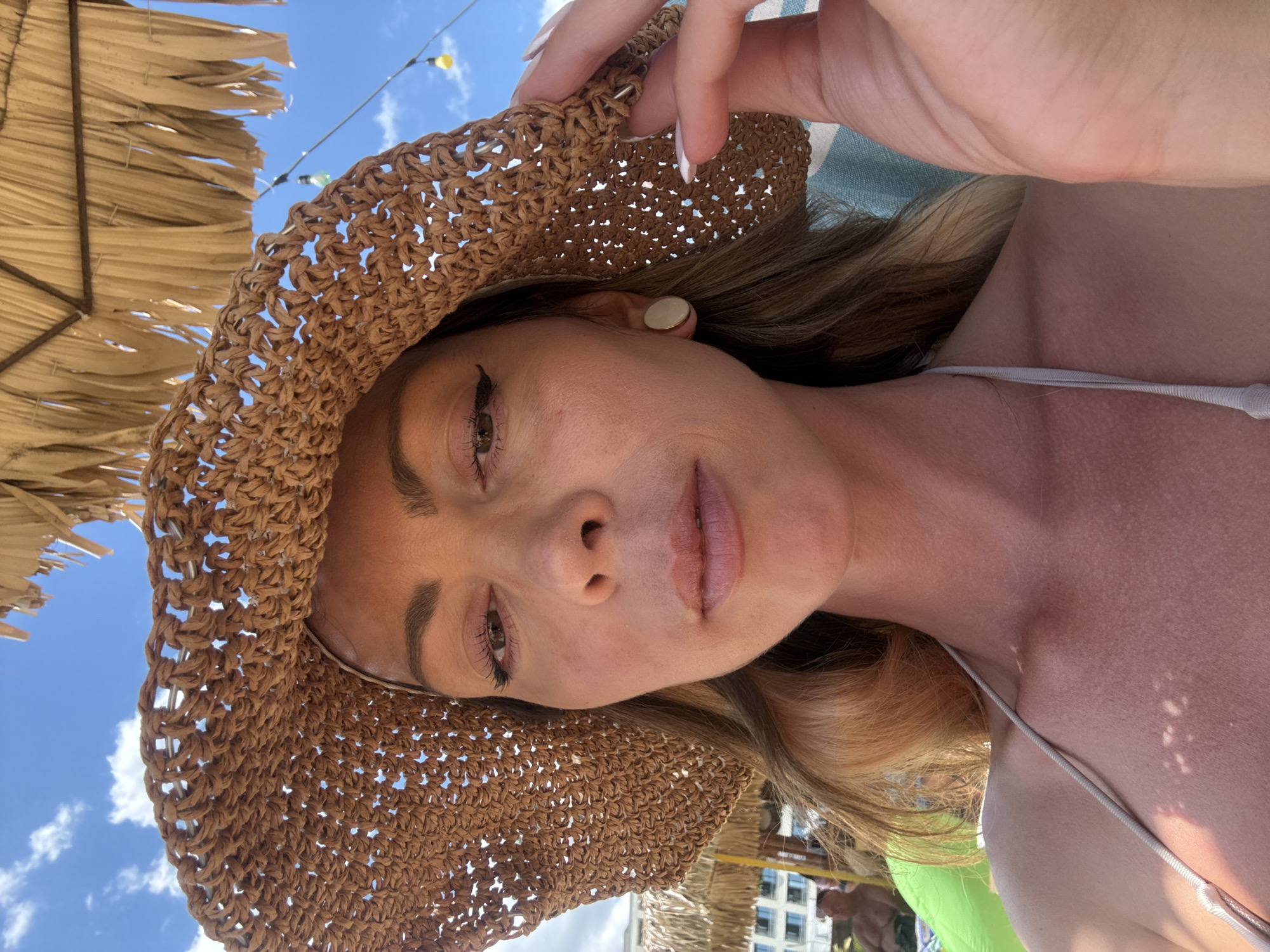 A medium shot selfie of a woman wearing a wide-brimmed straw hat and a bikini top on a sunny day. The woman has blonde hair, is wearing makeup, and has a light-colored circular earring. She is holding the brim of her hat with her right hand, which has white nail polish. The background shows a blue sky with clouds, a thatched umbrella, and string lights. In the blurred background, there appears to be a beach resort with buildings and other people. The context of the city of Nesebar, Bulgaria, is not visually discernible from the image itself.