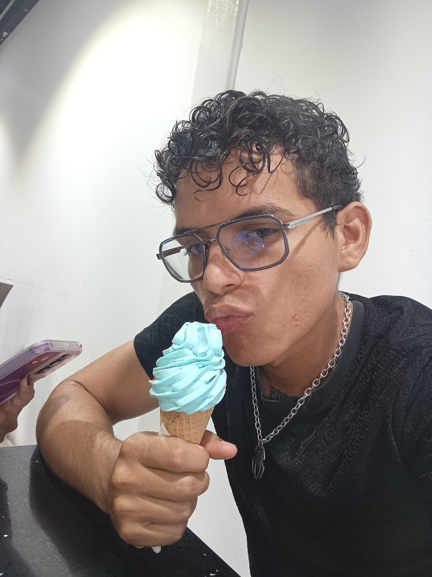 The image features a young man indoors, likely at a cafe or ice cream shop in Juan Griego, Venezuela. He is positioned at a dark, reflective counter against a plain white wall. The man has dark, curly hair and is wearing glasses with clear frames and dark temples. He is dressed in a dark, possibly black, textured short-sleeved top and accessorized with a silver chain necklace that has a dark, abstract pendant.
He holds a light blue ice cream cone in his right hand. The ice cream is a single scoop of a vibrant, pastel blue flavor in a classic waffle cone. The man's lips are puckered in a playful "kissing" or "duck face" expression, directed towards the ice cream, suggesting a humorous pose for the photograph.
To the left side of the frame, partially visible, is another person's hand holding a purple smartphone encased in a clear cover. The setting is illuminated by artificial light, creating a bright atmosphere. The overall mood is casual and lighthearted. There is no clearly visible text in the image.