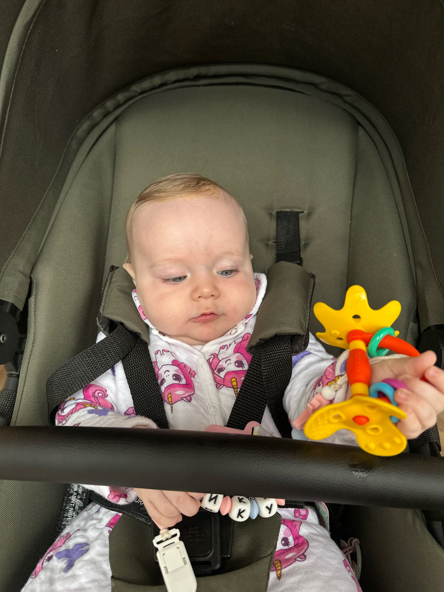 A baby is sitting in a dark green stroller, wearing a white onesie with a pink unicorn pattern. The baby has light skin and light hair, and appears to be looking down at a colorful toy in its hand. The toy is yellow with red and blue rings. The stroller has black safety straps. The image appears to have been taken indoors or in a shaded outdoor area. There is no indication of the location being Florianópolis, Brazil.