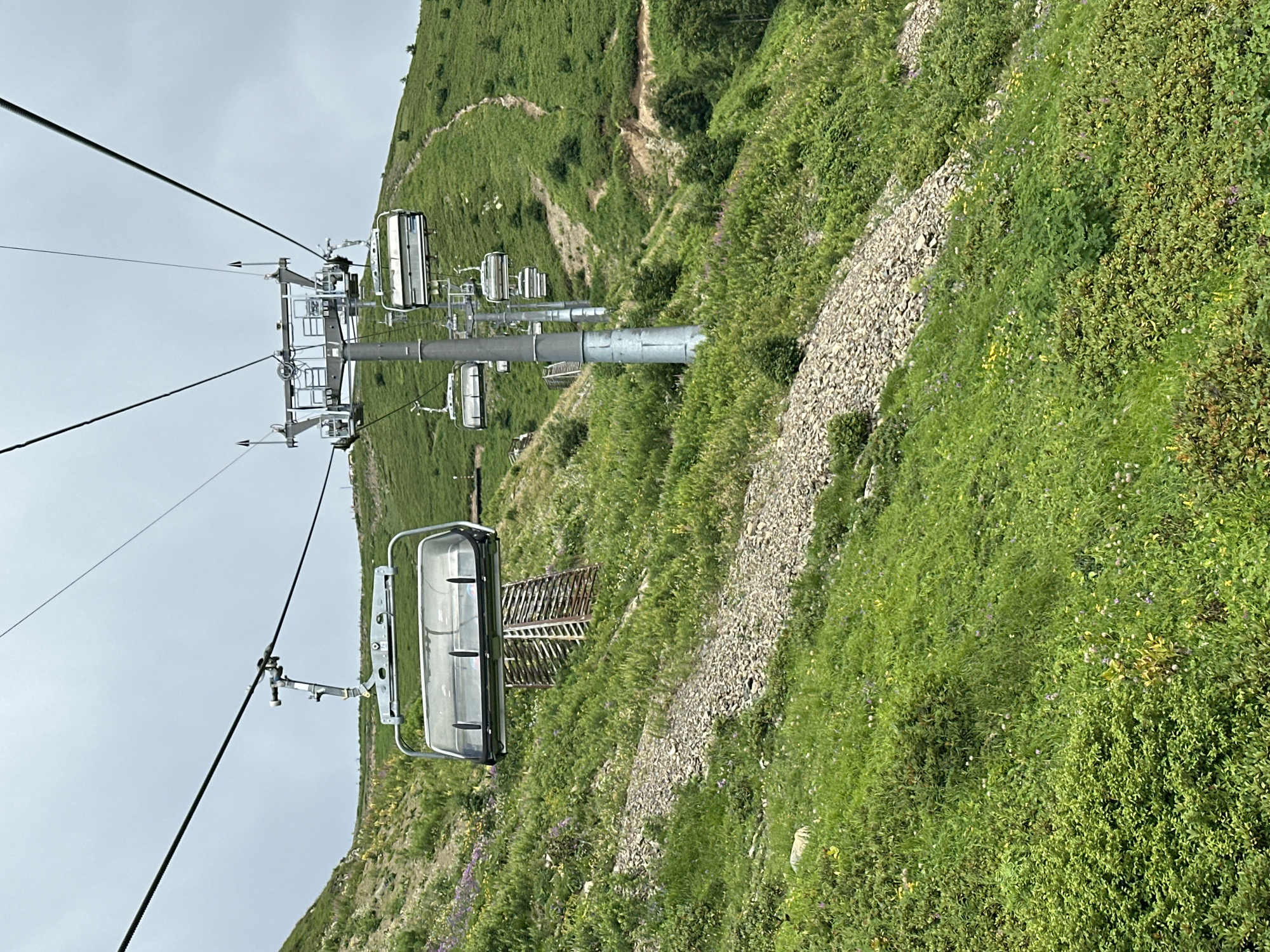 This image captures a ski lift ascending a lush, green mountainside in Estosadok, Russia. The sky is overcast, suggesting a cloudy day. The ski lift consists of multiple chairs suspended from a cable, with one chair prominently in the foreground. The mountainside is covered in vibrant green grass and dotted with scattered rocks and small purple and yellow wildflowers. A faint, winding path is visible on the upper part of the mountain. The scene is devoid of people, emphasizing the natural landscape and the infrastructure of the ski resort.
