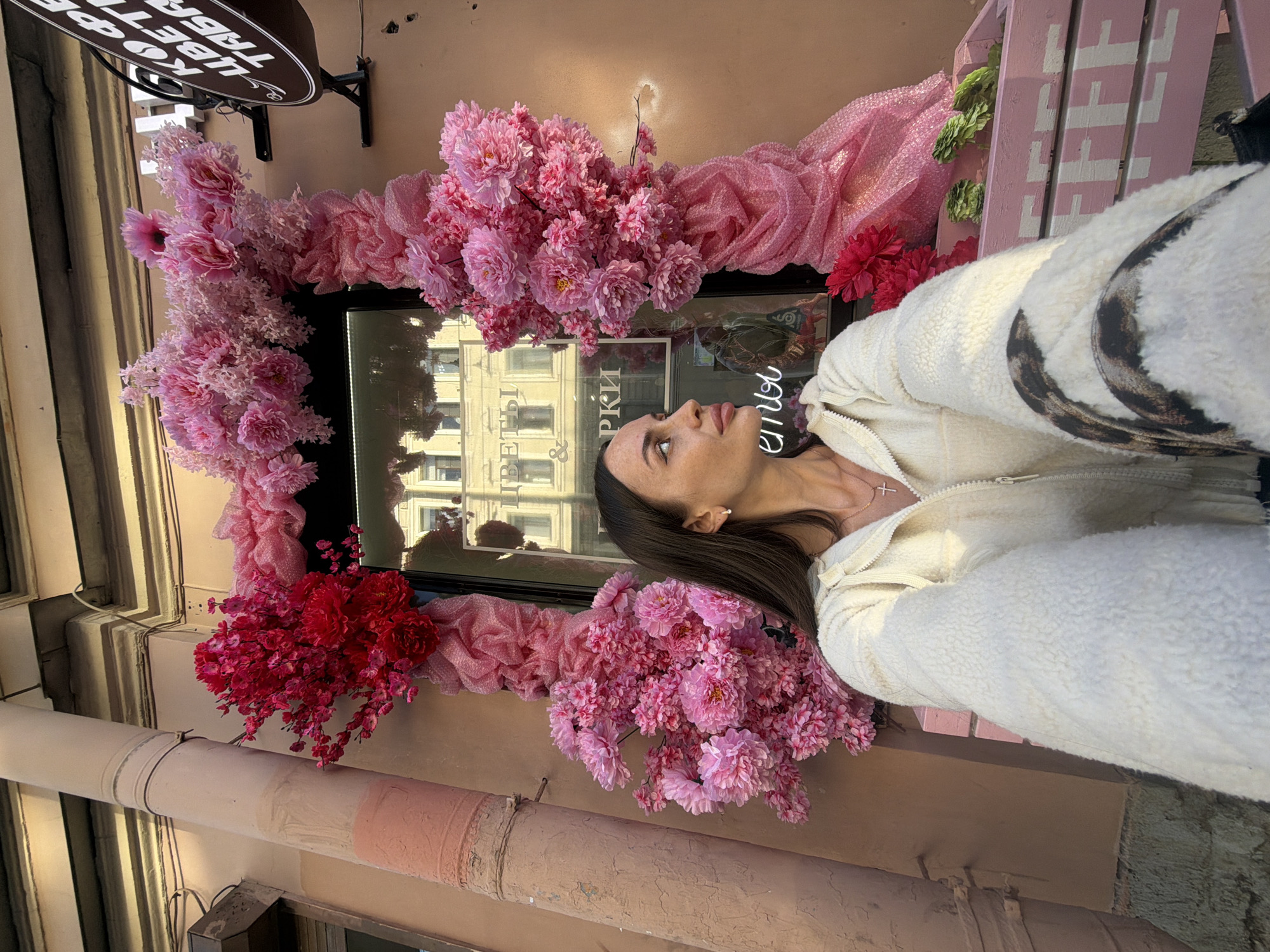 A woman with dark hair and freckles is taking a selfie outdoors in front of a storefront decorated with an abundance of pink and red flowers. The storefront appears to be a flower shop, with a window displaying a building reflection. A sign with Russian text is visible to the right of the entrance. The woman is wearing a cream-colored fleece jacket and a necklace with a cross. A pink planter box with green flowers sits in front of the storefront. The architectural style suggests the location is in Saint Petersburg, Russia.