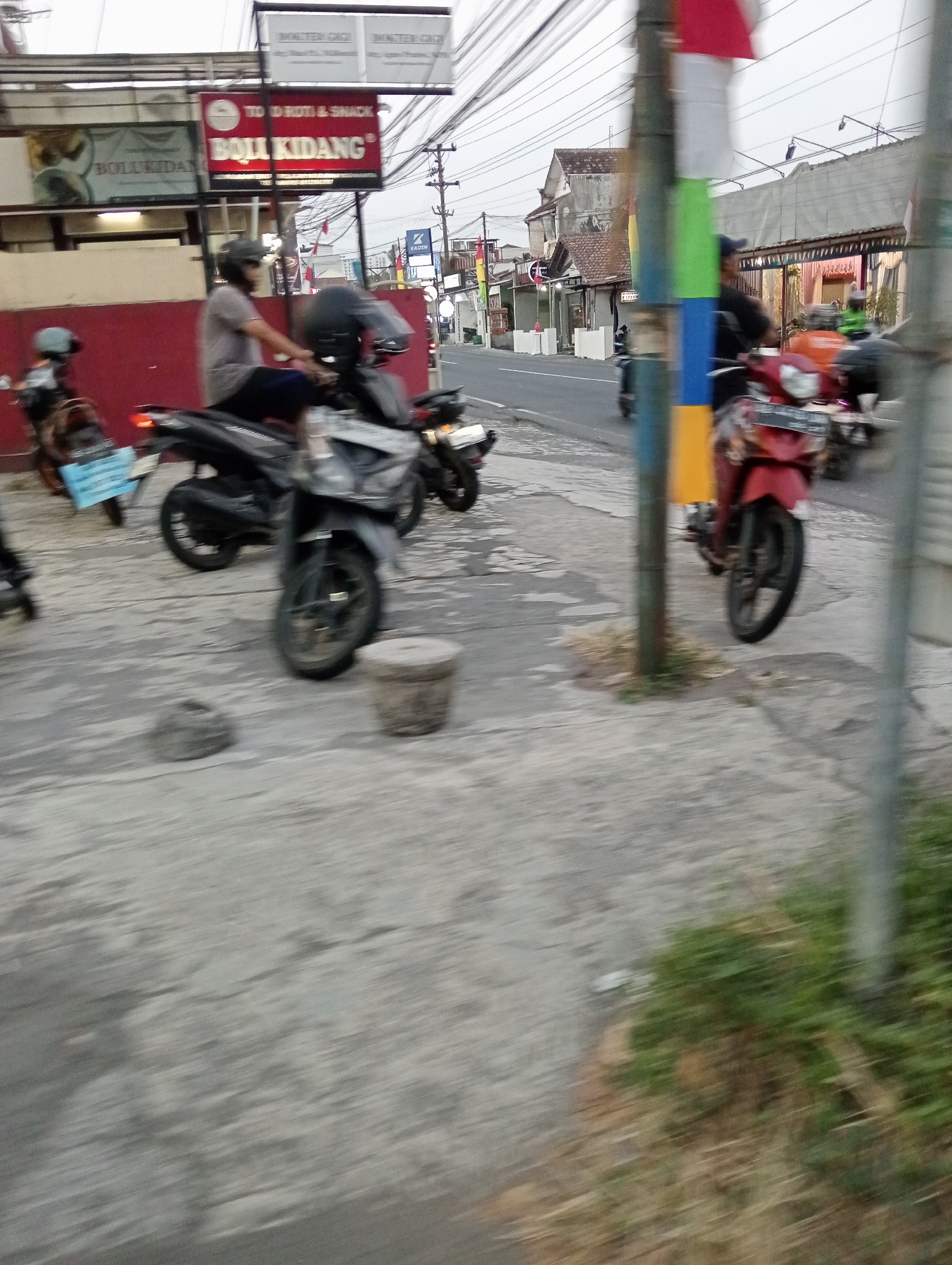 In Ngaglik, Indonesia, a street scene unfolds with several motorcycles parked and in motion on a paved sidewalk and roadway. A person wearing a helmet and a grey shirt is seated on a black motorcycle, facing left. Another motorcycle, black with silver accents, is parked in front of the first. A red motorcycle with a license plate is in motion to the right, and a white and blue motorcycle is parked on the left. In the background, several buildings are visible, including one with a red sign that reads "BOLU KIDANG" and another with a blue sign. Utility poles and overhead wires crisscross the sky. The overall environment appears to be an urban street with commercial establishments.