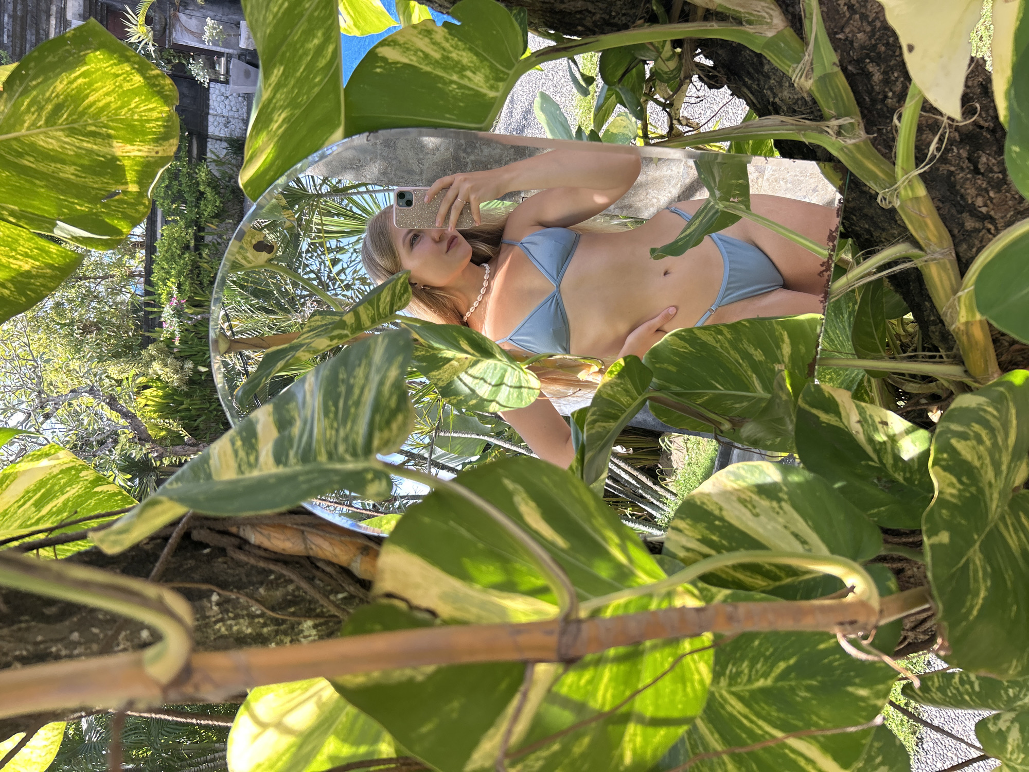 A woman in a blue bikini is reflected in a round mirror surrounded by leafy foliage, likely pothos plants, attached to a tree. The woman is holding a smartphone and appears to be taking a selfie. The environment suggests an outdoor setting, possibly a garden or natural area. The city of Kuta Selatan, Indonesia is not discernable from the available details.