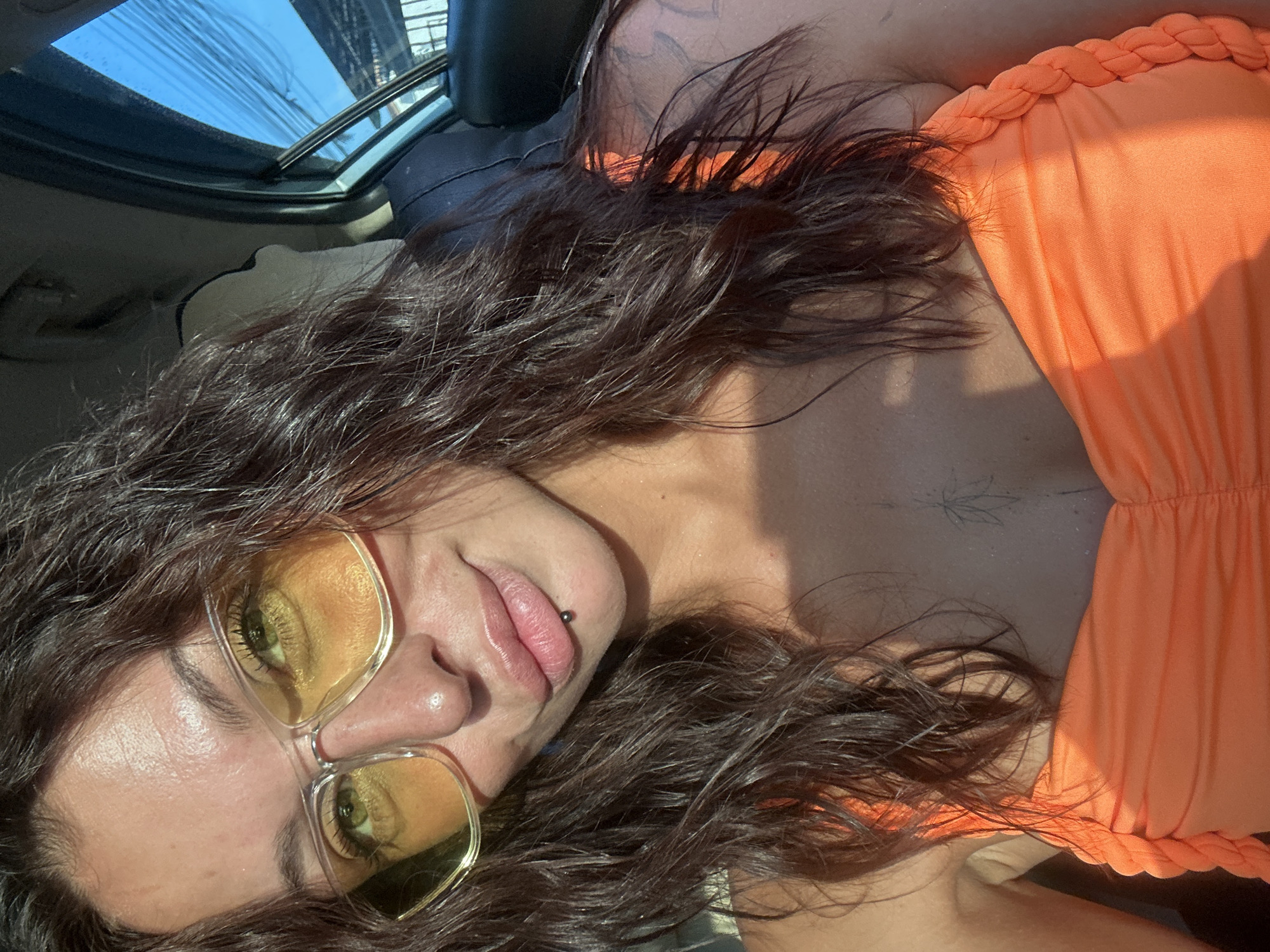 A woman with dark brown wavy hair, wearing large yellow-tinted aviator glasses and an orange top, is captured in a close-up selfie inside a vehicle. She has a lip piercing and a faint tattoo on her upper chest. The background shows the interior of a car, with a window reflecting bright blue sky and what appears to be outdoor structures. The lighting suggests it is daytime, with sunlight casting shadows on her face and clothing. The location is identified as Ko Samui District, Thailand.