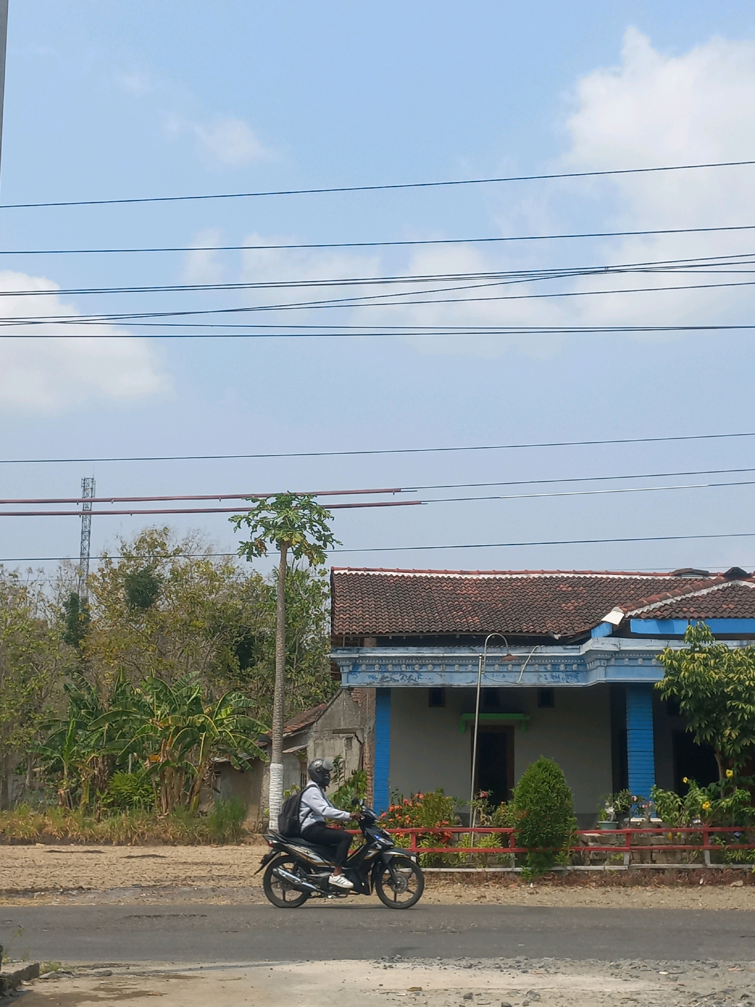 A person wearing a helmet and a backpack rides a black motorcycle on an asphalt road. They are seen from the side, moving from left to right. In the background, there is a house with a red-tiled roof and blue accents. Several trees and some vegetation are present around the house. Overhead, multiple power lines stretch across the frame. The sky is light blue with some scattered clouds. The location is Baturetno, Indonesia.