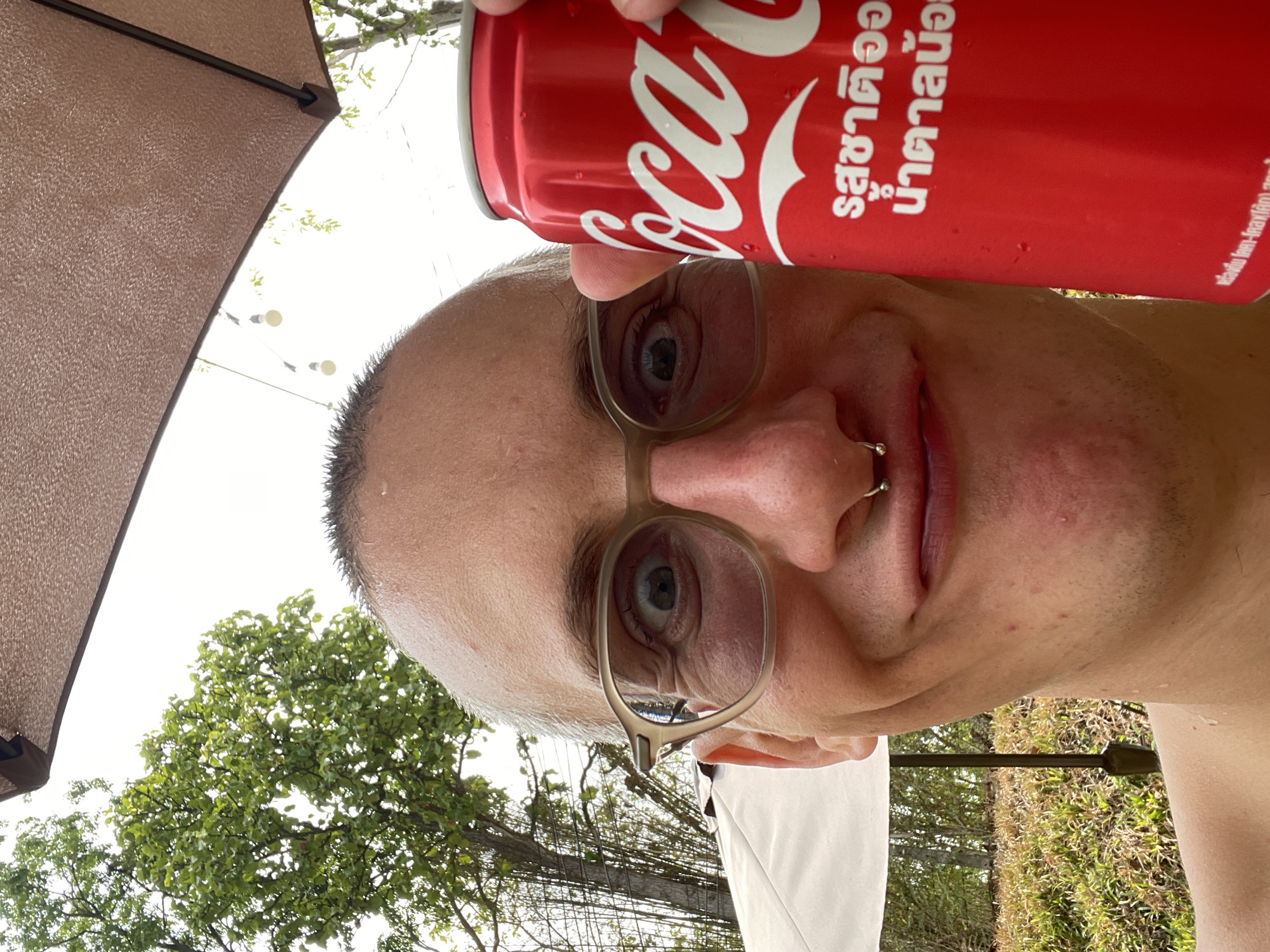 A close-up, low-angle shot captures a person holding a red Coca-Cola can next to their face. The person has light skin, short, wet hair, and wears glasses. A nose ring is visible. In the background, partially obscured by an umbrella, are trees and foliage. The location is Tambon Nam Phrae, Thailand, based on the text on the Coke can.