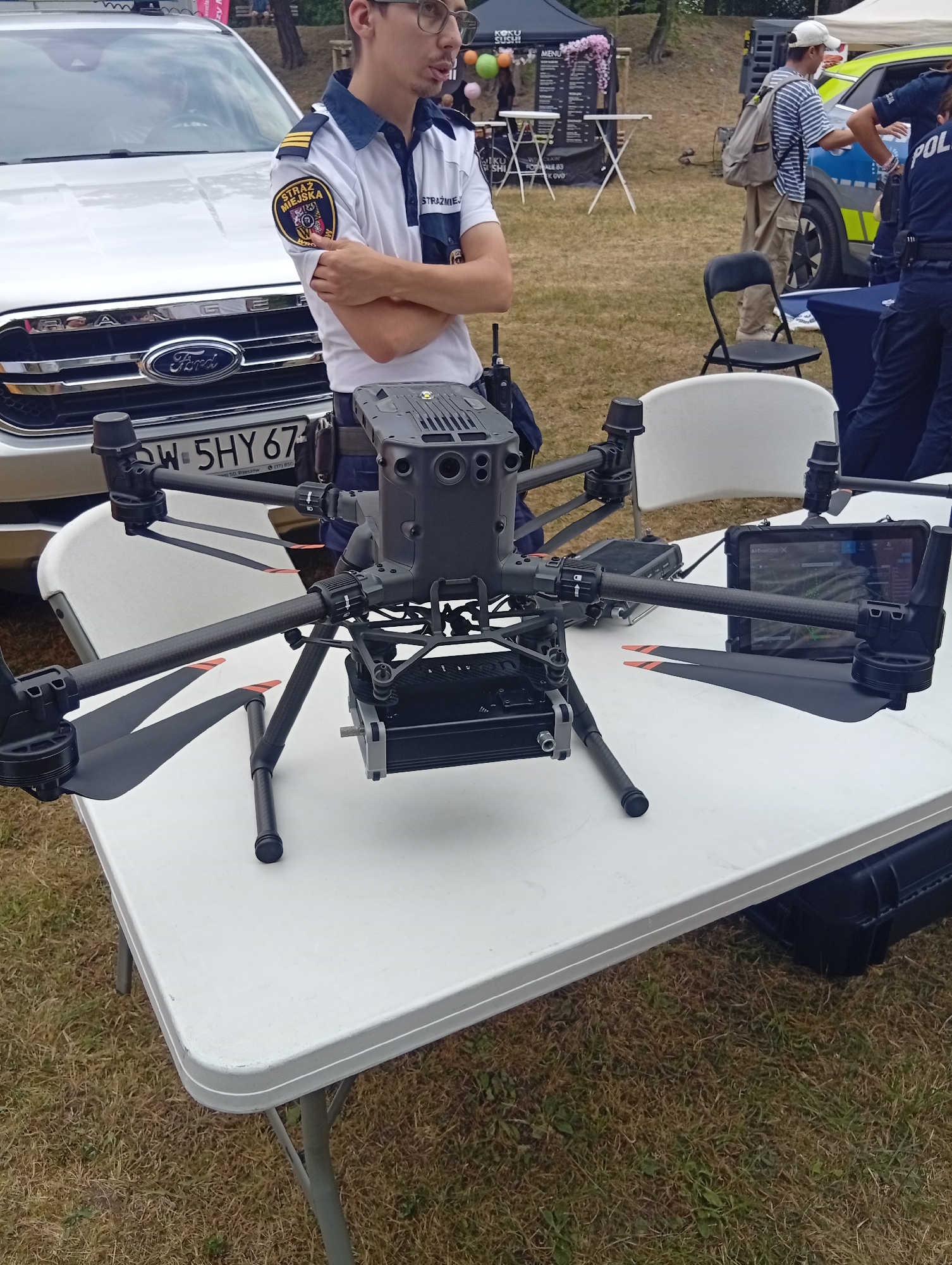 A man in a white shirt with a "STRAŻ MIEJSKA" (City Guard) patch and navy blue collar and epaulets stands near a silver Ford Ranger pickup truck. He has his arms crossed and is wearing glasses. In front of him, on a white folding table, is a large black drone with its propellers extended. Behind him, a person in a striped shirt and cap walks away, and a person in a police uniform stands by a white and blue police car. The scene appears to be outdoors on a grassy area with some tents and tables in the background. The license plate on the Ford Ranger reads "DW 5HY67". Based on the license plate format and the presence of the "STRAŻ MIEJSKA" and police, this scene is likely in Poland. Given the specific detail of the license plate format, this observation is reinforced. While the exact location in Wrocław, Poland cannot be definitively determined from the image alone, the general context suggests an event or demonstration likely held in a public space within the city.