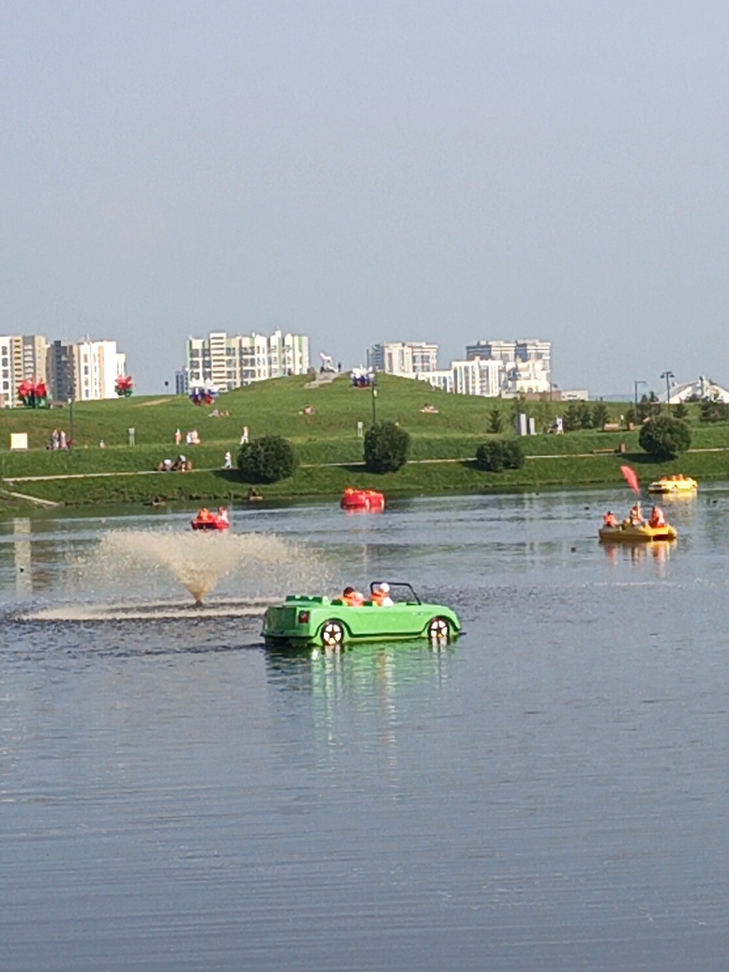 This image depicts a park scene with a lake and contemporary buildings in the background. In the foreground, several colorful paddle boats are on the water. One green paddle boat shaped like a car, with two people inside, is prominently featured on the right side. Further out on the lake, a yellow paddle boat with a red flag and three people is visible. Another red paddle boat is also present in the mid-ground. A fountain is active on the left side of the lake, creating a spray of water. The background consists of a grassy hill dotted with people and trees, with a cluster of modern buildings behind it. This scene is characteristic of the "Park of the Replications" (Парк "Реплика") in Yekaterinburg, Russia, known for its artificial hills and recreational areas.