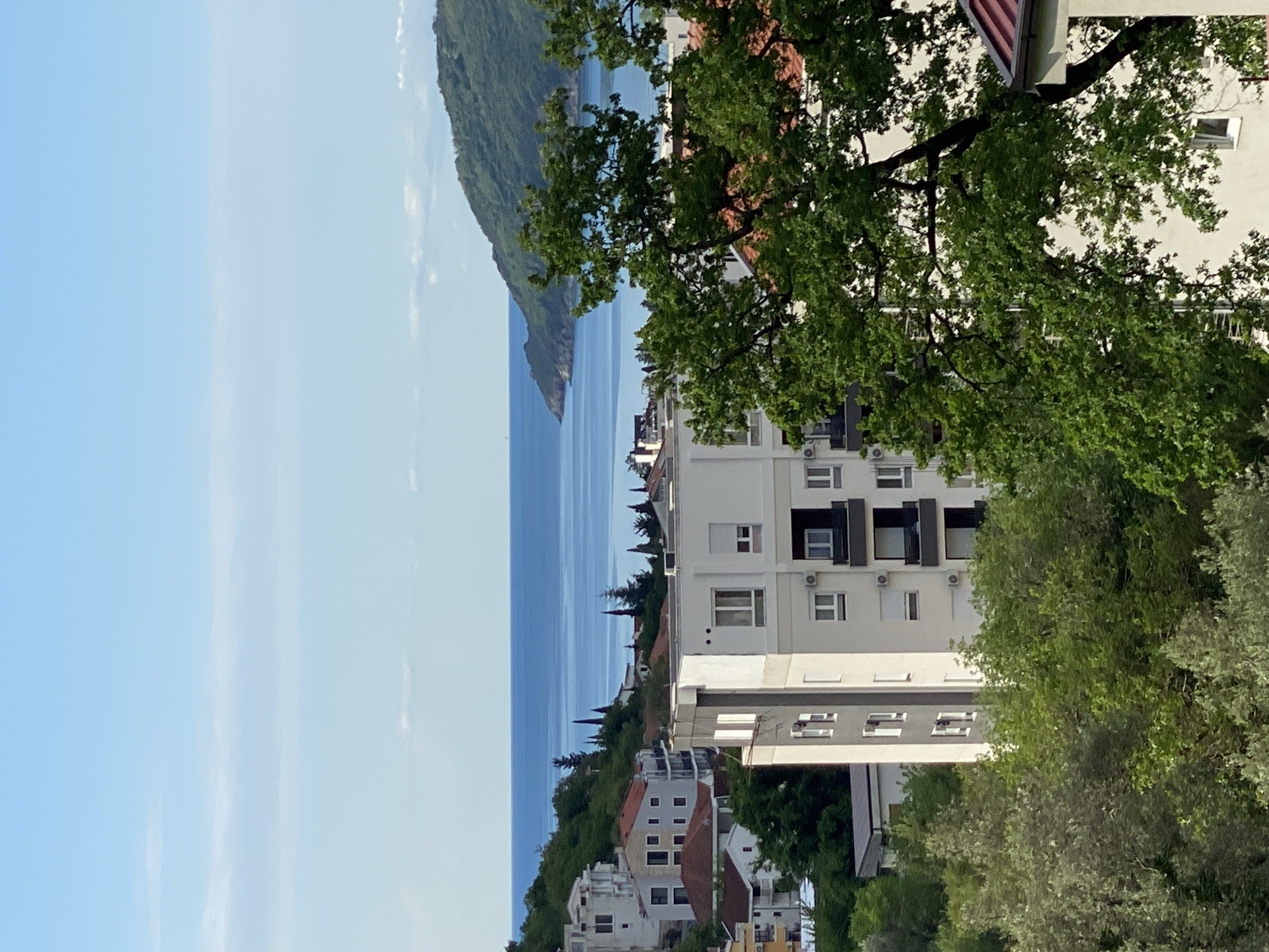 The image shows a view of the city of Budva, Montenegro, looking out towards the Adriatic Sea. In the foreground are several buildings, primarily white apartment blocks, interspersed with trees and vegetation. The architecture is modern, with multiple floors and balconies. The sea is visible in the middle ground, stretching out towards a mountainous peninsula that forms part of the coastline. The sky is light blue with wispy clouds. The image appears to be taken from an elevated position.