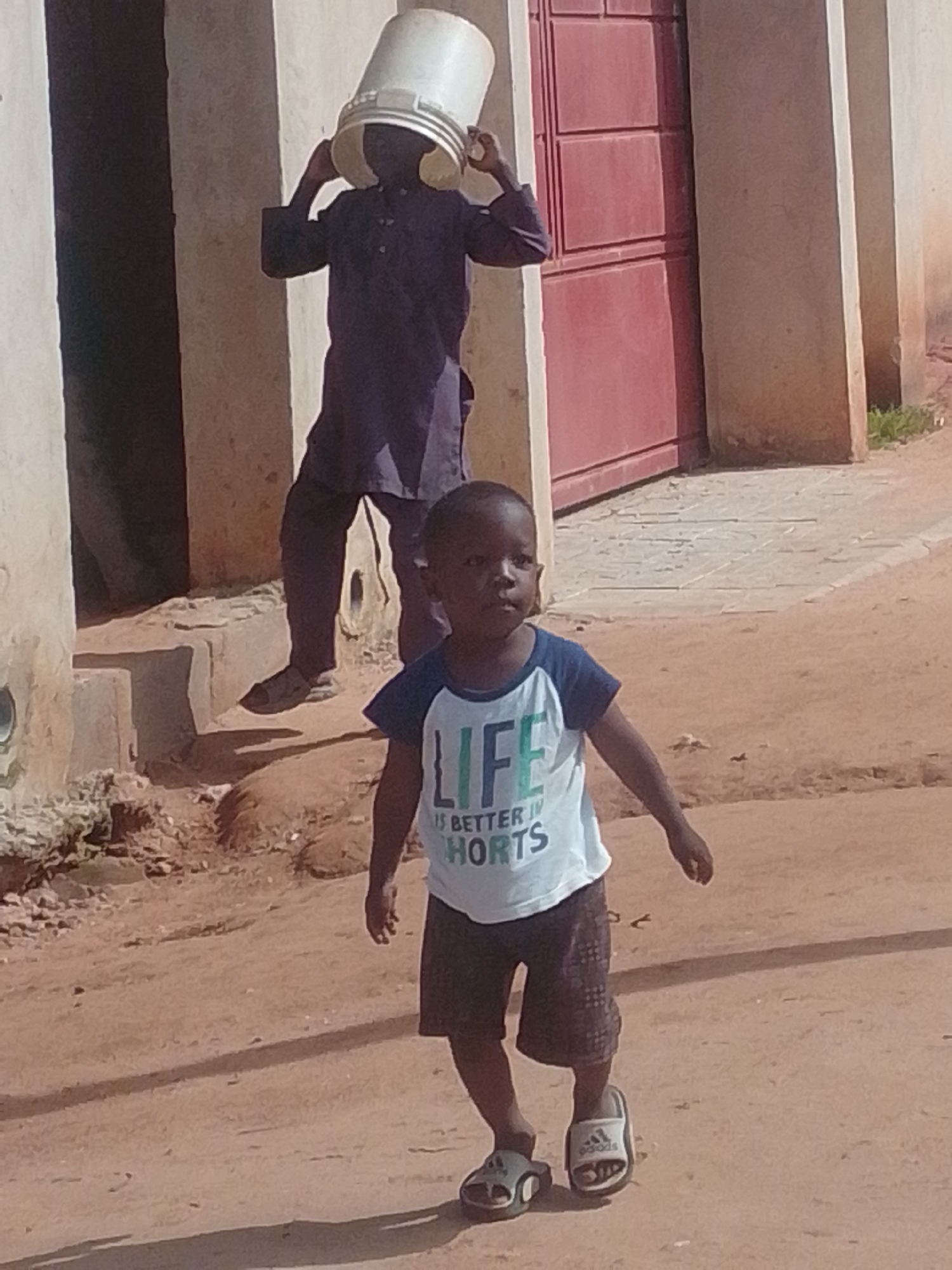 In an outdoor, dusty, urban environment, a young child walks towards the camera. The child is wearing a white t-shirt with blue sleeves and a graphic print on the front that reads "LIFE IS BETTER IN SHORTS". The child is also wearing dark-colored shorts and slip-on sandals. To the left and behind the child, an older person, wearing dark clothing and a bucket on their head, is walking away from the camera. The background includes a building with a red door and a textured wall. The ground is composed of dirt and sand, with some rocks and debris scattered about. The lighting suggests it is daytime with direct sunlight creating shadows.