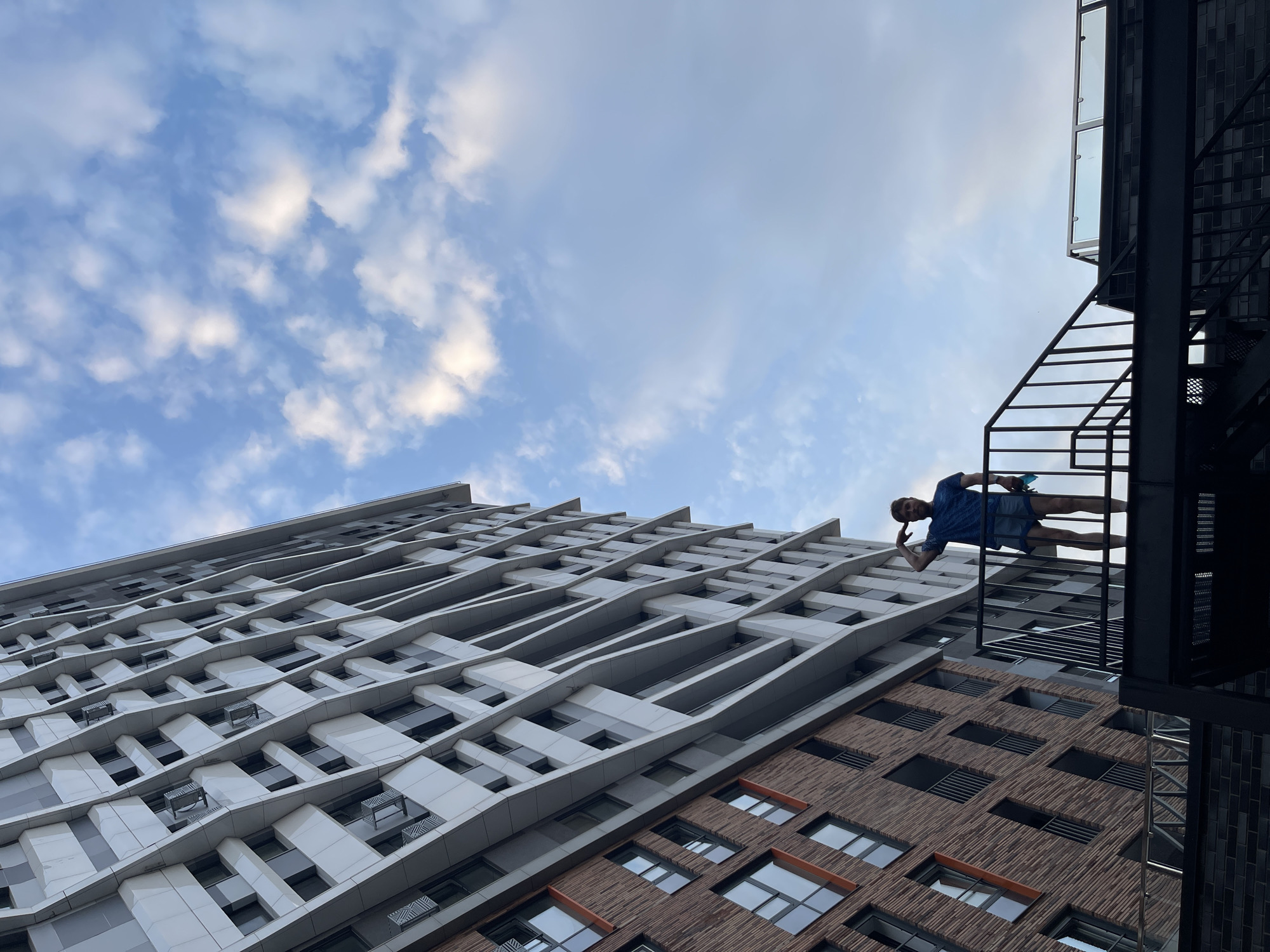 A low-angle shot captures a modern building with a distinctive geometric facade, predominantly white panels with grey accents, contrasted with a lower section clad in reddish-brown brick. A man, dressed in a blue t-shirt and denim shorts, stands on a black metal staircase in the foreground, saluting. The sky above is a pale blue with scattered white clouds, suggesting daytime. The architecture suggests an urban setting, possibly residential, with balconies and windows visible on the building. The scene appears to be an exterior view, possibly a rooftop or elevated walkway.