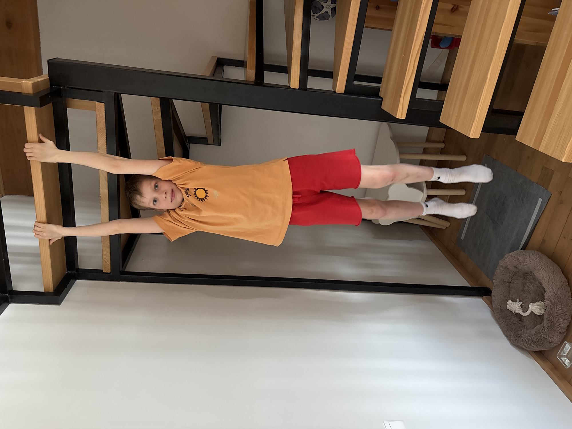 A child is suspended between the wooden beams of a bed frame and a ladder in a room. The child wears an orange t-shirt with a sun design, red shorts, and white socks. The bed frame has black metal supports. A gray mat and a dog bed are on the floor nearby. The location is unknown.