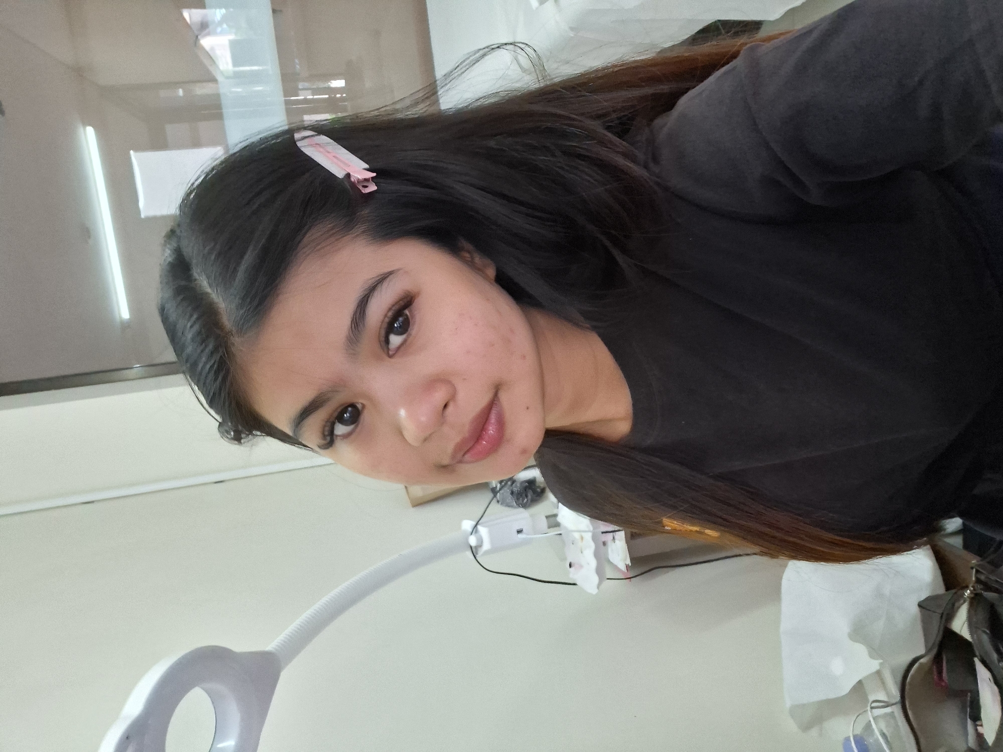 A young woman is pictured in a selfie taken in what appears to be a beauty salon in Kuta, Indonesia. She has long, dark hair with a pink and white hair clip securing a section on the right side. Her eyes are open, and she is looking at the camera with a slight smile. She is wearing a dark t-shirt. Behind her, a white magnifying lamp with a flexible neck is visible, along with a small table with some items on it. The background includes a wall and a blurred reflection of fluorescent lighting.