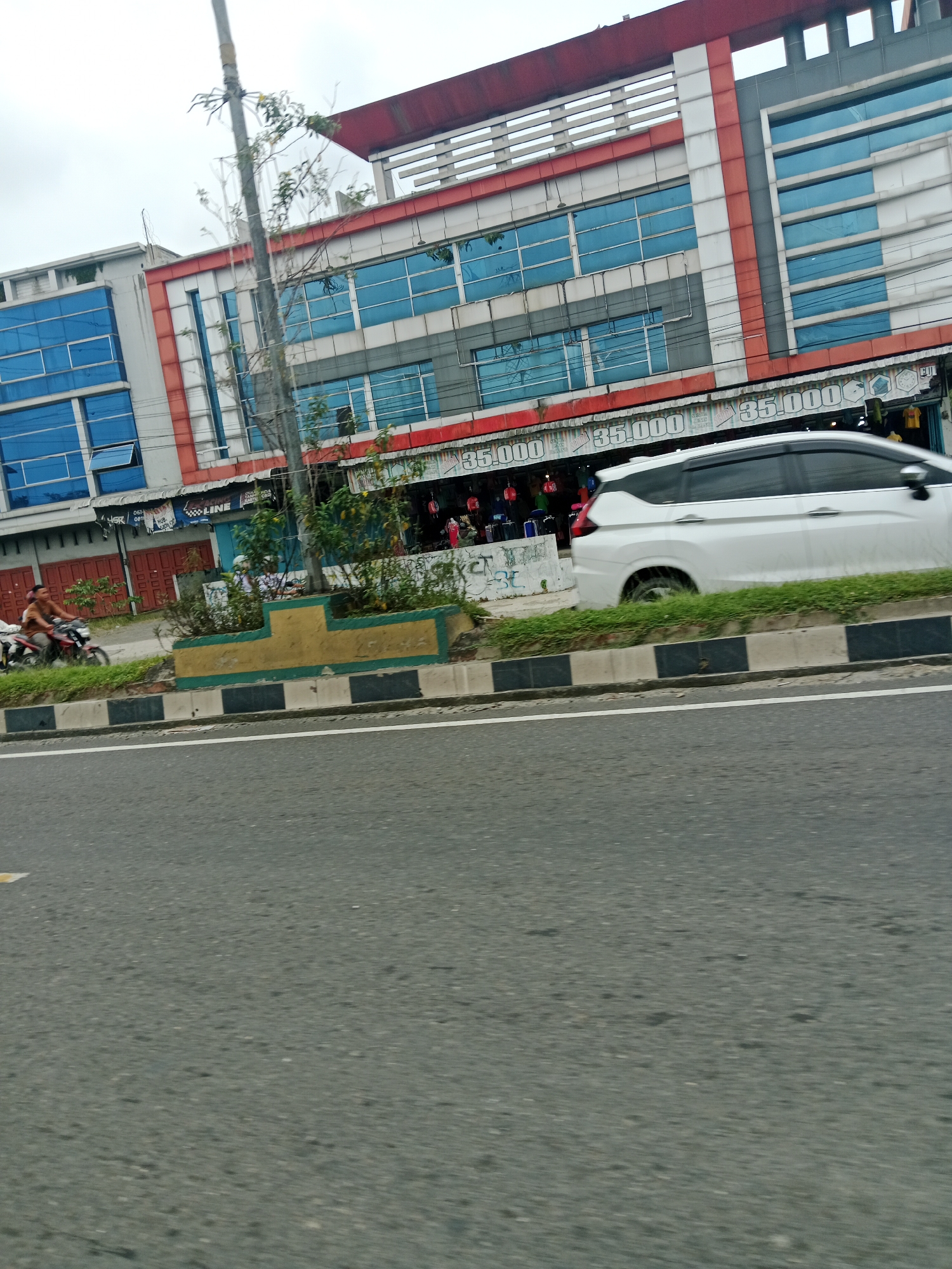A low-angle, slightly blurred, medium shot captures a commercial building and street scene in Binjai, Indonesia. The building is a multi-story structure with prominent blue glass windows and red and white accents. The ground floor appears to house shops, with signage indicating prices of "35.000" and various merchandise displayed. A white minivan is parked or moving in front of the building, partially obscuring the shops. To the left, a man on a motorcycle is visible. A concrete divider with yellow and green detailing separates the street from the building's frontage, and a thin tree grows in the foreground. The overall environment suggests a bustling urban commercial area on an overcast day.