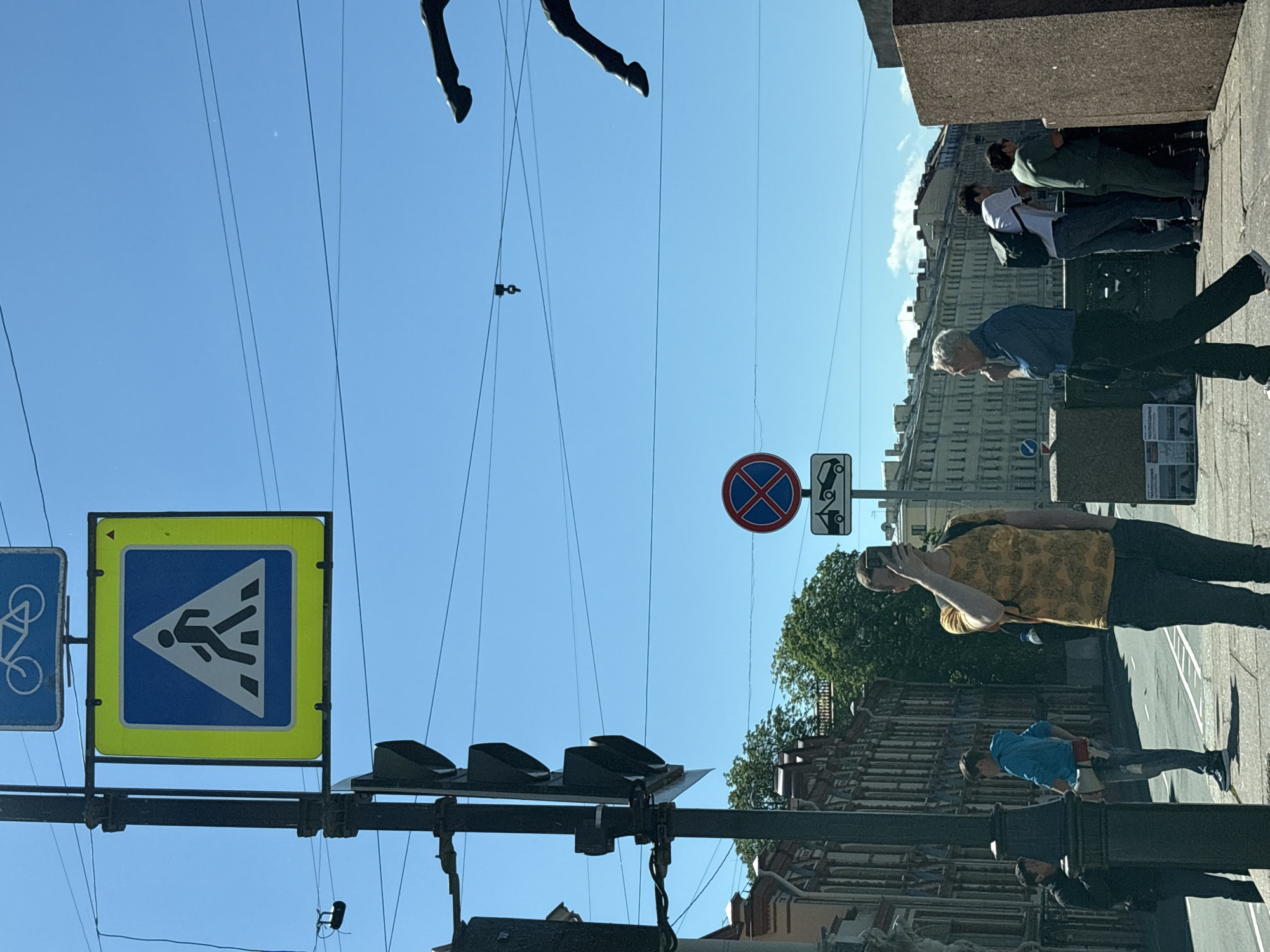 The image depicts a street scene in Saint Petersburg, Russia, under a clear blue sky. In the foreground, a person wearing a yellow t-shirt with a floral pattern stands facing away from the camera, holding a smartphone to their face. They have a backpack on. To their left, a man in a blue shirt and black pants walks past, and further left, two men are visible, one in a black jacket and another in a blue hoodie holding a megaphone. A traffic light pole with multiple signal lights is prominent on the left. Above the traffic light, a square yellow sign with a blue border displays a pedestrian crossing symbol. Further in the background, across the street, is a building with ornate architecture, typical of Saint Petersburg. Several other traffic signs are visible, including a circular red and blue sign indicating no stopping, and a rectangular sign below it depicting a tow truck. The background also shows more buildings and trees. A portion of a statue or sculpture, appearing to be horse legs, is visible in the upper right corner of the frame, suggesting the location might be near a bridge or monument. The overall scene conveys a sense of urban life with people going about their day.