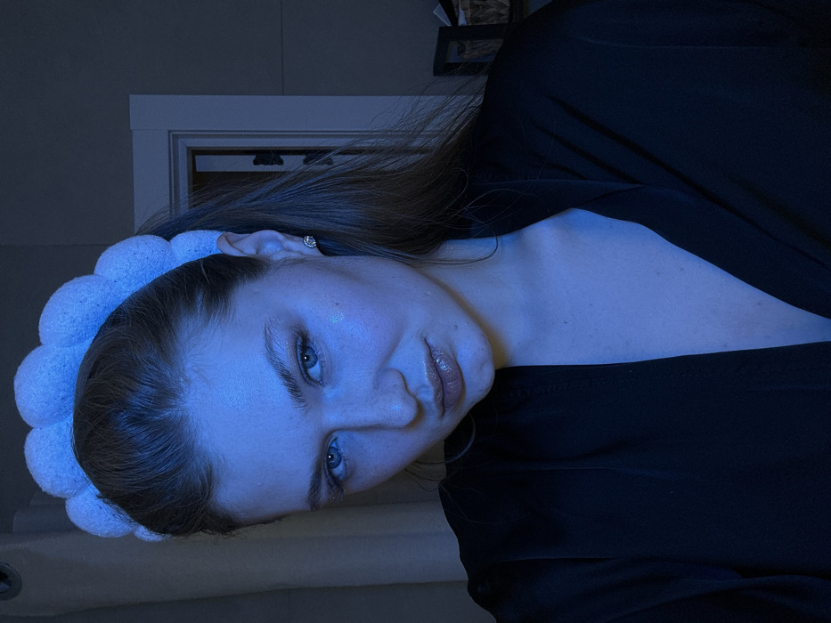 A woman with light skin, blue eyes, and light brown hair is wearing a white headband and a black top. The lighting is cool and bluish, giving the scene a somewhat subdued atmosphere. The woman is positioned at an angle, with a slight tilt of her head. The background features a doorway. Her expression is neutral. An earring is visible.