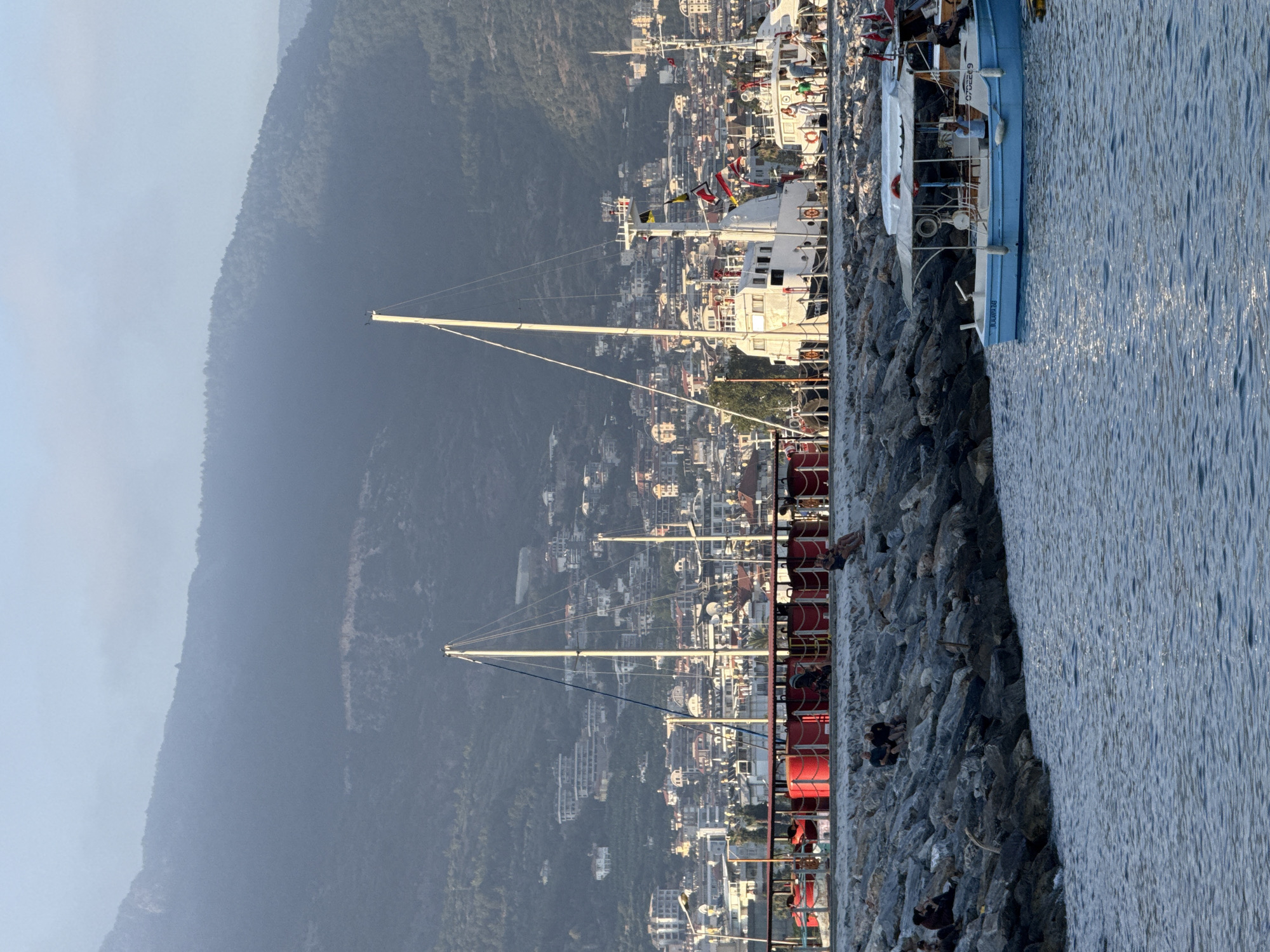 The image shows a town nestled against a steep, forested mountain in Alanya, Turkey. The town is characterized by white buildings with red tile roofs, indicative of the region. The foreground is dominated by a rocky breakwater or pier, with several boats docked alongside. One boat, a blue and white vessel, is clearly visible, with people on deck. Another larger white boat with masts and flags is also prominent, alongside a red-hulled boat. The water is calm and reflects the light, suggesting a clear day. The time of day appears to be late afternoon or early evening, given the soft lighting and long shadows. Several people are visible on the rocky area, some sitting and others standing, enjoying the view or the boats. The overall atmosphere is peaceful and scenic, with a hint of coastal life and tourism. The text "BLU WIN 38" is visible on the blue boat.