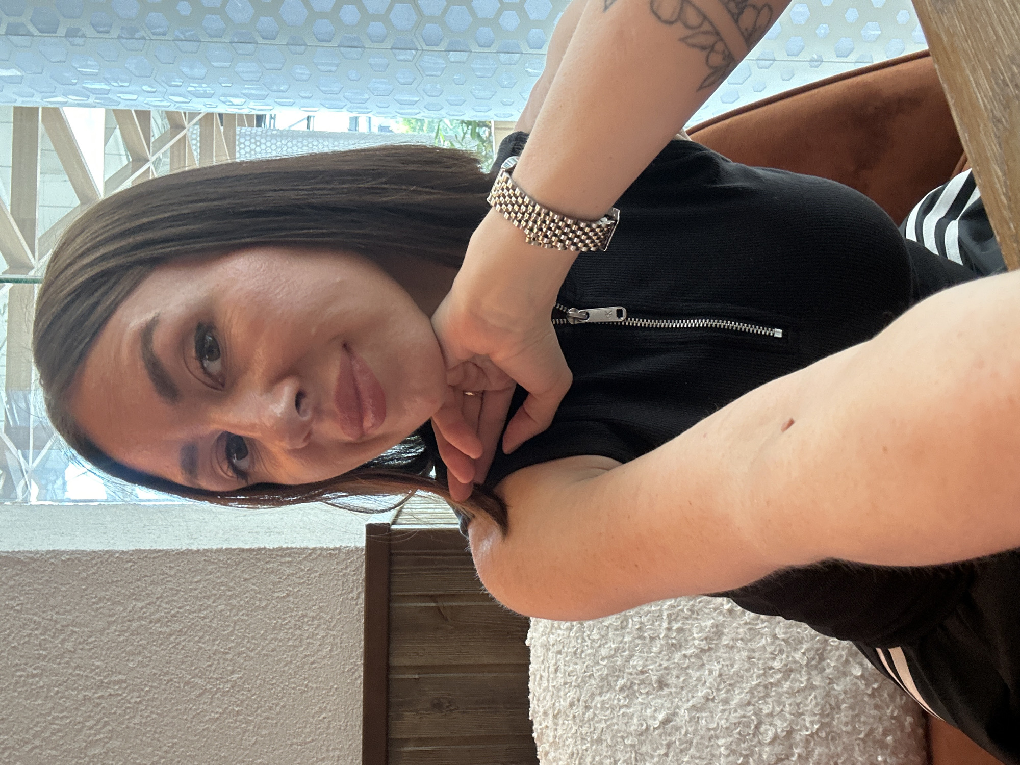 A medium-close-up photo shows a woman from the chest up, looking directly at the camera with a slight smile. She has long, dark brown hair and is wearing a black top with a prominent silver zipper down the front. Her right hand is positioned near her chin and neck, with her fingers intertwined, while her left arm is bent, revealing a tattoo on her upper arm. On her right wrist, she wears a textured, two-tone (silver and gold) metallic bracelet. A small mole is visible on her right forearm.
The background suggests an indoor setting, likely during the daytime due to bright, diffused light. Behind the woman, a large window or ceiling feature with a light-colored hexagonal pattern is visible. Below this, wooden architectural elements or furniture can be seen. To the right, part of a brown, upholstered chair or sofa is present. The lower part of the image shows a textured white or light gray surface, possibly a wall or fuzzy fabric. No specific text is visible in the image. The scene is set in Kazan, Russia.