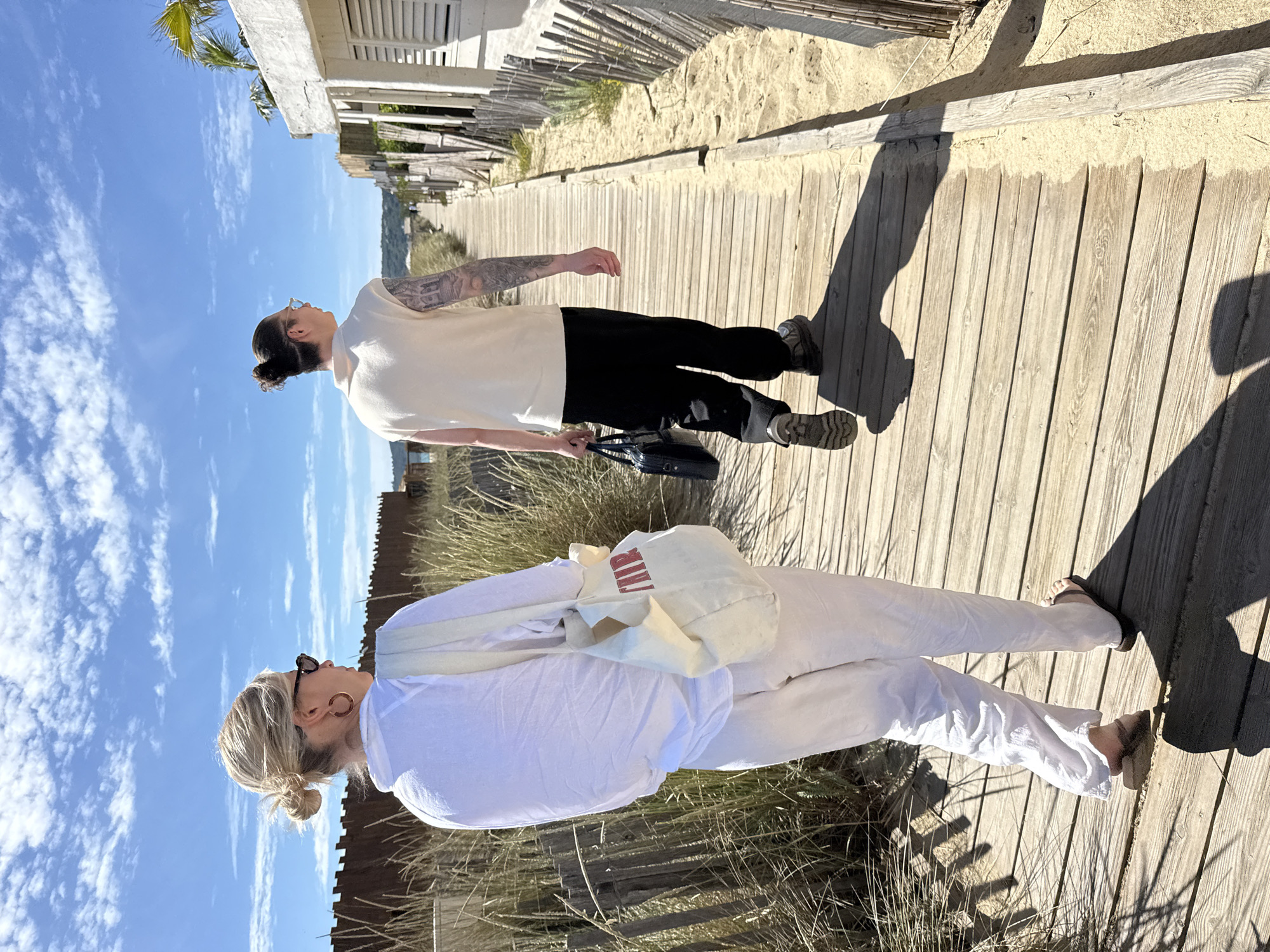 Two people are walking on a wooden boardwalk, potentially near a beach.
One person, on the left, is wearing a white outfit, including pants, a top, and a shoulder bag. They also have sunglasses and their hair pulled back. The other person is wearing a cream-colored top and black pants, holding a black bag.
The boardwalk is surrounded by sand and vegetation. A fence runs along the side of the boardwalk. In the background, there is a glimpse of buildings. Based on the location and details, the scene is likely in or near Ramatuelle, France.