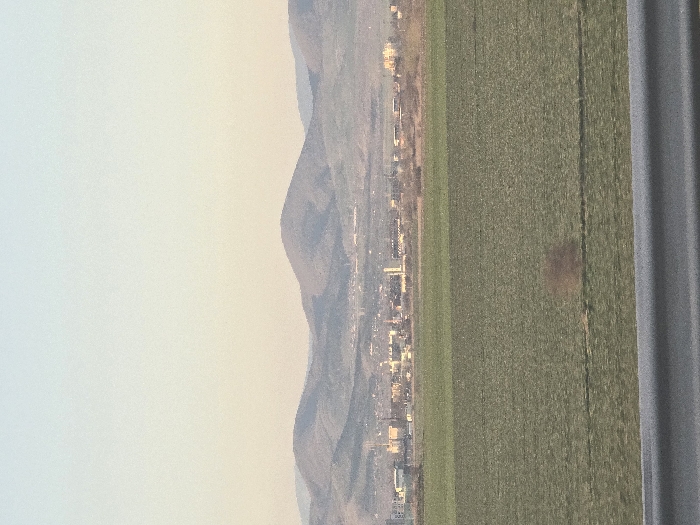 The image depicts a panoramic landscape overlooking the city of Diadovo, Bulgaria, under natural light conditions. In the immediate foreground, a blurred gray horizontal metal barrier, consistent with a guardrail, spans the bottom of the frame. Above this barrier, an expansive, flat, green agricultural field occupies the midground, extending towards the distant city. Faint horizontal striations are visible within the field, suggesting cultivation. A diffused, brownish, indistinct form is present in the lower-central portion of the field, possibly a reflection or distant vegetation.
Beyond the field, the intermediate background reveals the cityscape of Diadovo, characterized by a cluster of industrial and commercial buildings. Several structures exhibit prominent vertical elements, such as chimneys or towers, indicative of industrial activity. The city appears somewhat hazy due to distance.
The background consists of a series of rolling, dark gray-blue mountains or hills that rise above the city and extend across the horizon. The sky, occupying the upper portion of the frame, displays a subtle gradient, transitioning from a pale blue at the zenith to a light yellowish-orange hue near the mountain peaks, consistent with ambient daylight. No individuals, explicit actions, or interactions are discernible within the frame.