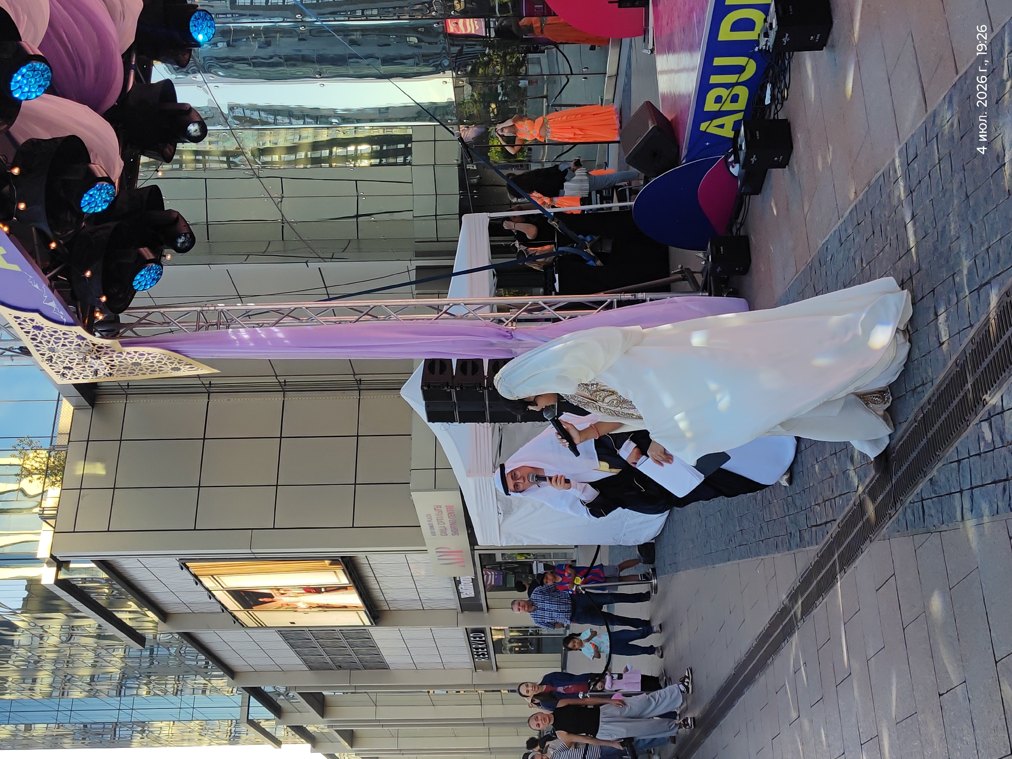 The image captures an outdoor event taking place on a sunny day in Astana, Kazakhstan, likely on July 4, 2026, at 7:26 PM. The setting appears to be a pedestrian area in front of a modern building with a glass facade. A stage is set up with lighting equipment, purple and white drapes, and a banner that reads "ABU DI." Two individuals are standing at the front of the stage, holding microphones and appearing to be presenting or performing. One is wearing traditional Arabic attire, including a white headscarf and a dark robe, and the other is wearing a white robe with ornate detailing.
Behind the stage, several people are gathered, observing the event. Among them are women and children, some dressed casually. The building in the background features multiple stories with reflective glass windows, suggesting a commercial or office complex. Signs on the building indicate the presence of shops, with one prominently displaying the word "RESERVED." The ground is paved with stone tiles, and a drainage grate runs across the foreground. The overall atmosphere suggests a public gathering or festival.