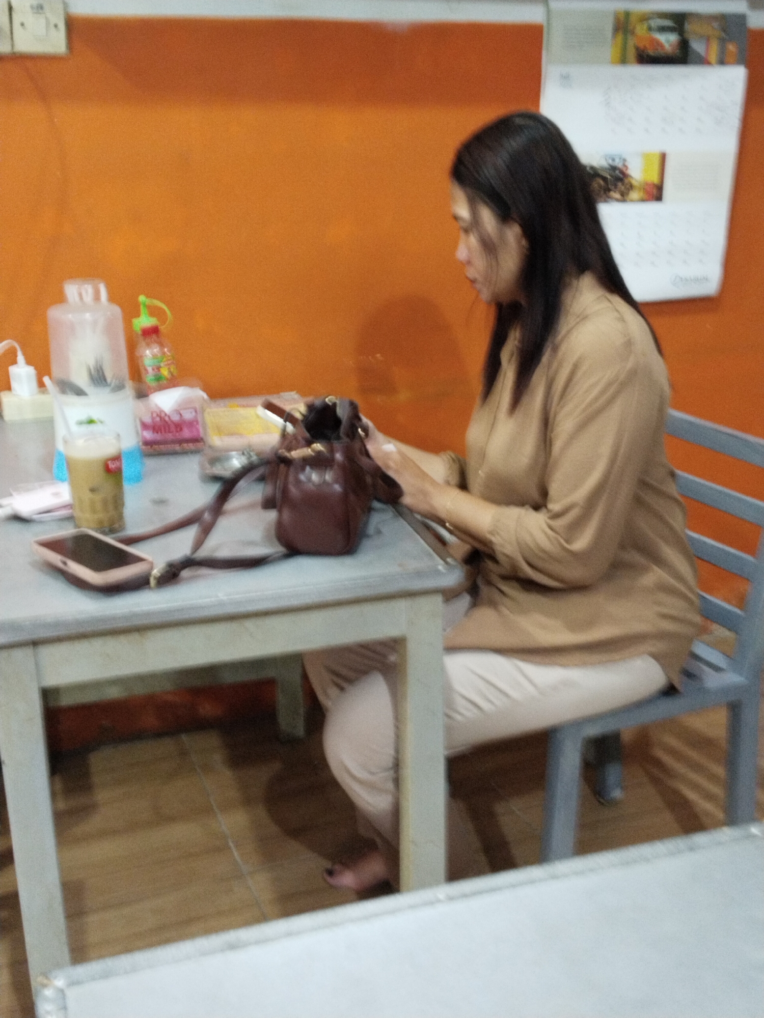 A woman in a light brown blouse and light-colored pants sits at a gray table in an indoor setting with an orange wall and wooden floor. On the table are a glass of iced coffee, a brown leather handbag, a mobile phone, a bottle of sauce, and what appears to be a packet of cigarettes. A calendar is visible on the wall behind the woman. The scene suggests a casual dining or waiting area, possibly a cafe or restaurant in Prigi, Indonesia.