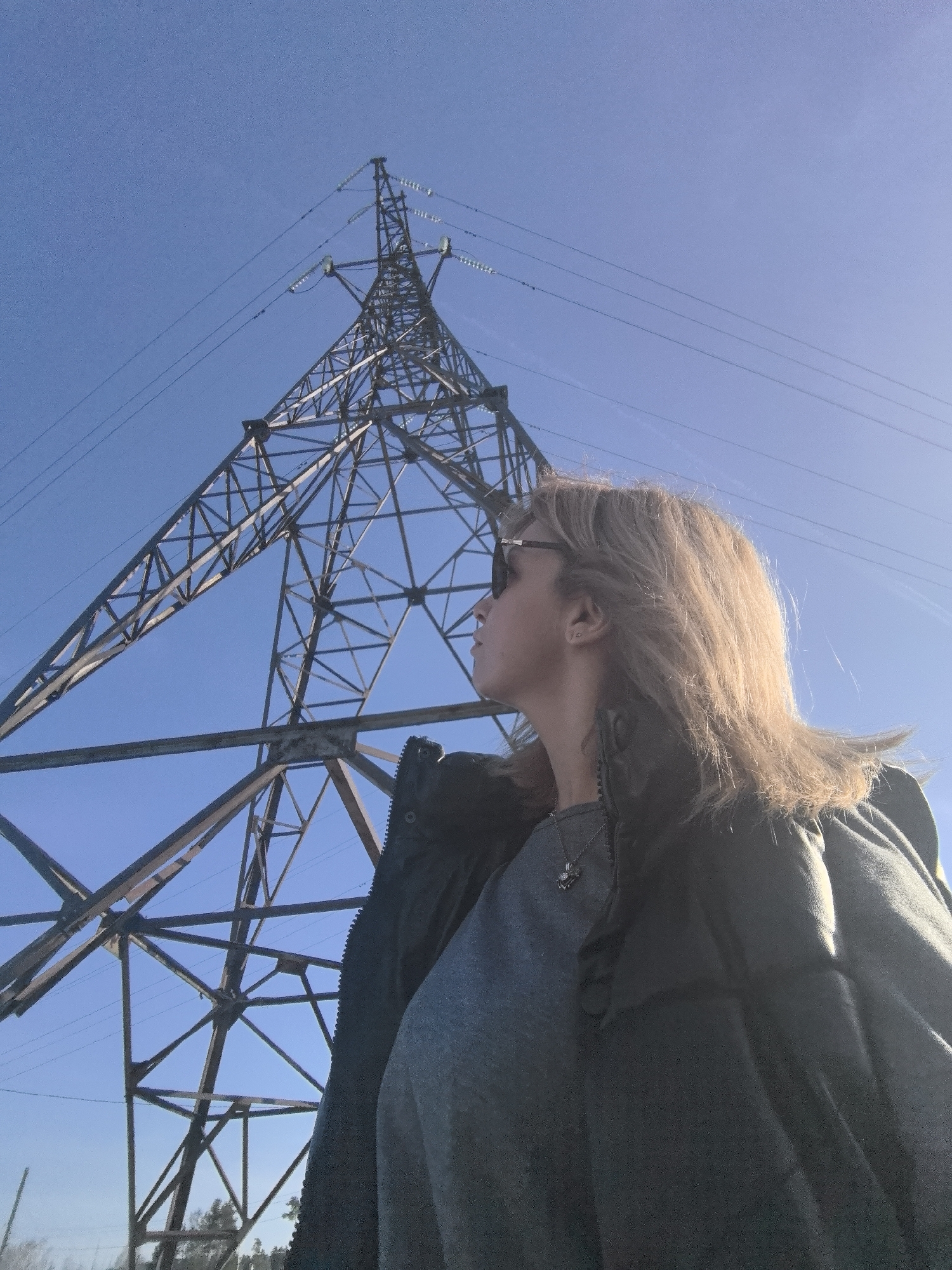 A medium, low-angle shot captures a woman standing beside an electricity pylon under a clear blue sky. The woman, positioned in the foreground, is wearing sunglasses, a gray shirt, and a dark-colored jacket or vest. Her gaze is directed upward, following the towering pylon. The pylon is made of a lattice of steel beams, forming a tall triangular structure that tapers towards the top. Electric wires are strung between the pylon's arms. The scene suggests an outdoor location, potentially on the outskirts of a city, given the industrial nature of the pylon. The location is in Noviy, Russia. The lighting indicates a sunny day, with shadows subtly defining the structure of the pylon and the contours of the woman's face and clothing.