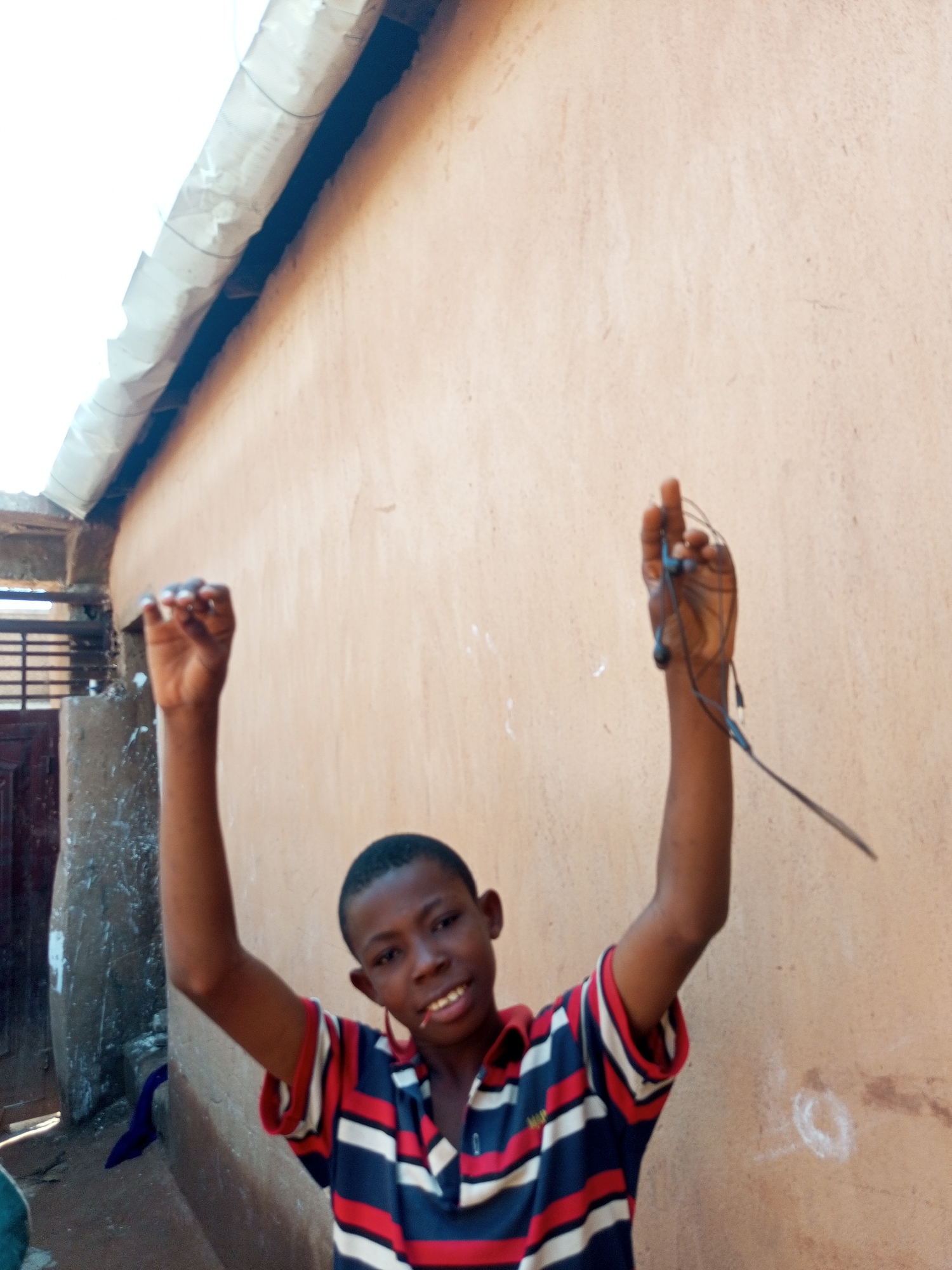 A young boy is standing outside a building with a peach-colored wall. He is wearing a blue, red, and white striped shirt. His arms are raised, and he is holding a pair of blue earbuds in his right hand. The boy is smiling and looking towards the camera. In the background, there is a white awning or roof extending from the building, and a dark doorway or opening to the left. The ground appears to be dirt or concrete. The overall scene suggests an outdoor residential or communal area in Tempure, Nigeria.