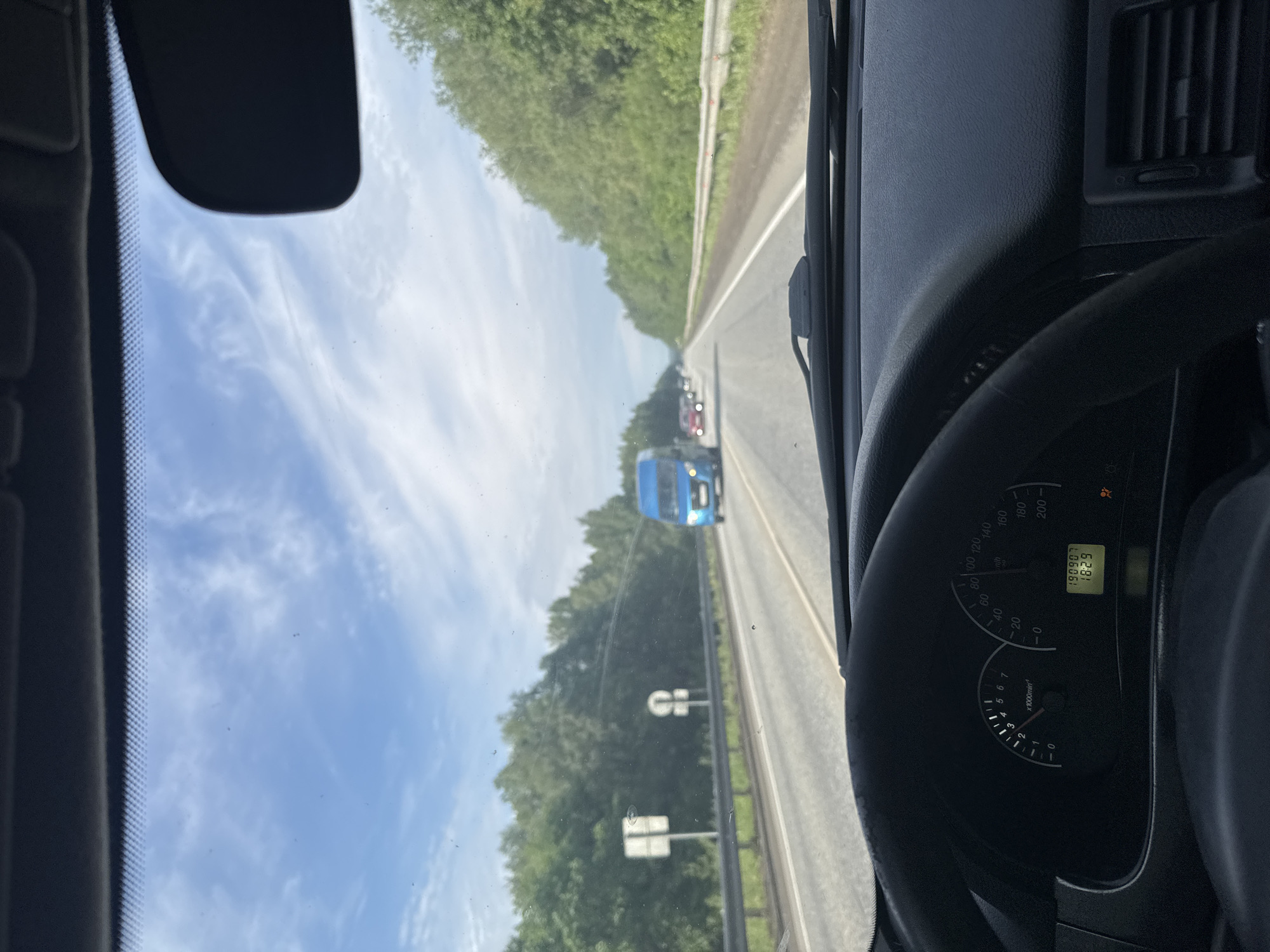 The image, taken from inside a vehicle, captures a driver's perspective of a road trip near Усть-Сыны, Russia. The scene looks forward through the windshield onto a highway bordered by dense green trees under a bright blue sky with scattered white clouds. The weather appears clear and sunny.
Ahead on the road, several vehicles are visible, including a prominent blue van, indicating light to moderate traffic. A road barrier runs along the right side of the highway. Partially visible road signs are too distant to read.
Inside the car, the right side of the frame shows a portion of the dashboard and the instrument cluster. The speedometer needle points to approximately 80-90 km/h, and the tachometer shows the engine running at around 2000-2500 RPM. A digital display on the cluster shows "190907", likely the odometer reading in kilometers, and "18:29", indicating the time is 6:29 PM. An orange warning light, possibly an airbag or seatbelt indicator, is illuminated on the cluster. A part of the rearview mirror is visible in the upper left corner of the frame. The overall impression is of a late afternoon or early evening drive on a highway through a forested area.