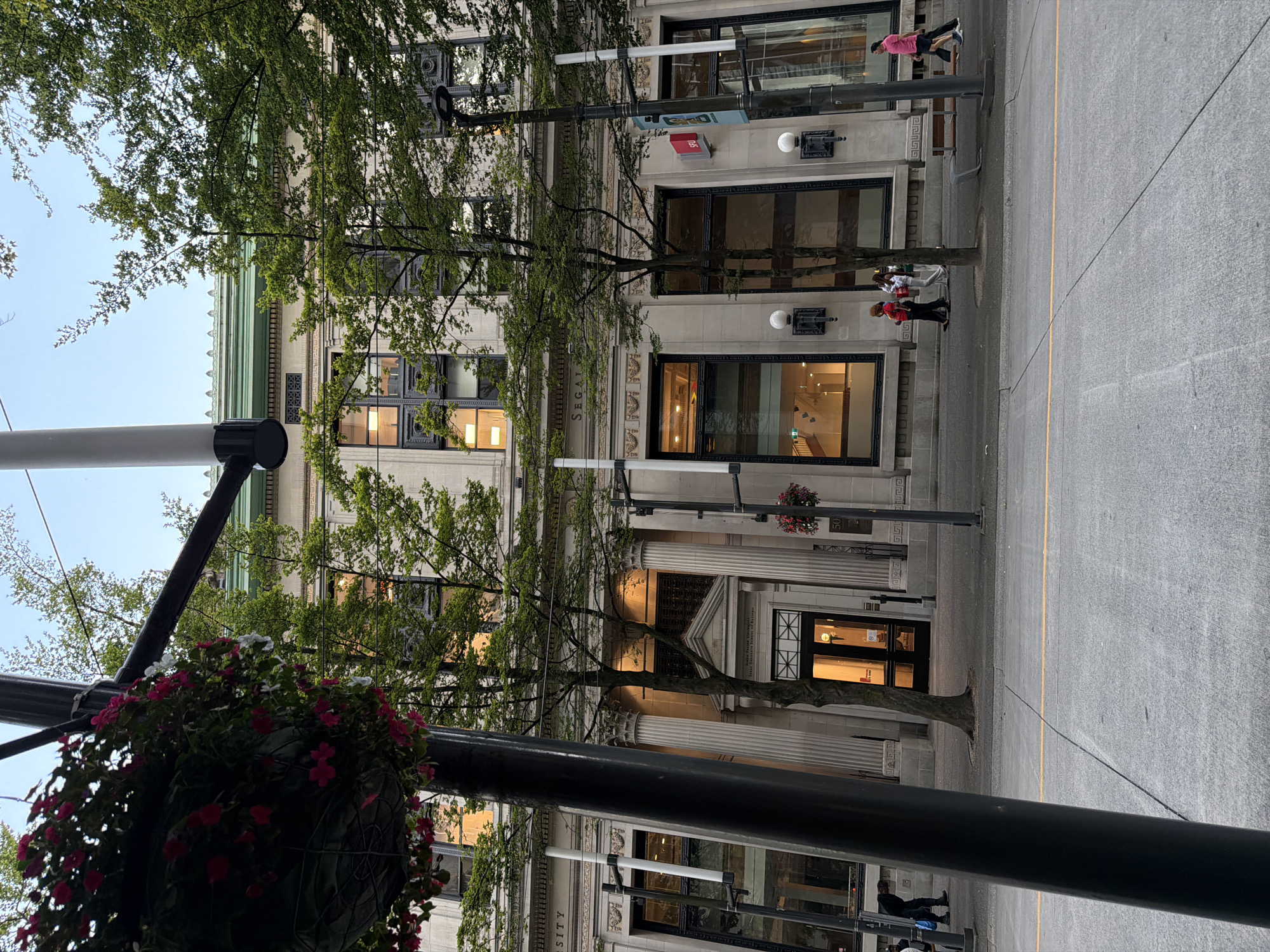 The image depicts a street scene in Vancouver, Canada, featuring a stately building with classical architectural elements and lush green trees lining the sidewalk. The building appears to be of significant historical or institutional importance, with large windows, stone facade, and ornate detailing. Several trees are prominently placed in front of the building, their branches reaching towards the structure and obscuring parts of the facade.
On the sidewalk in front of the building, there are several people walking. One individual in the foreground is partially obscured by a dark pole and hanging plants, but appears to be moving away from the camera. Further down the sidewalk, a group of three individuals are walking together, one person wearing a bright red top. To the right, another person in a pink shirt is walking. Streetlights and some decorative lighting fixtures are visible along the sidewalk. The street itself shows a yellow line marking. The overall atmosphere suggests an urban environment with a touch of nature, likely during the daytime or early evening, given the lighting conditions.