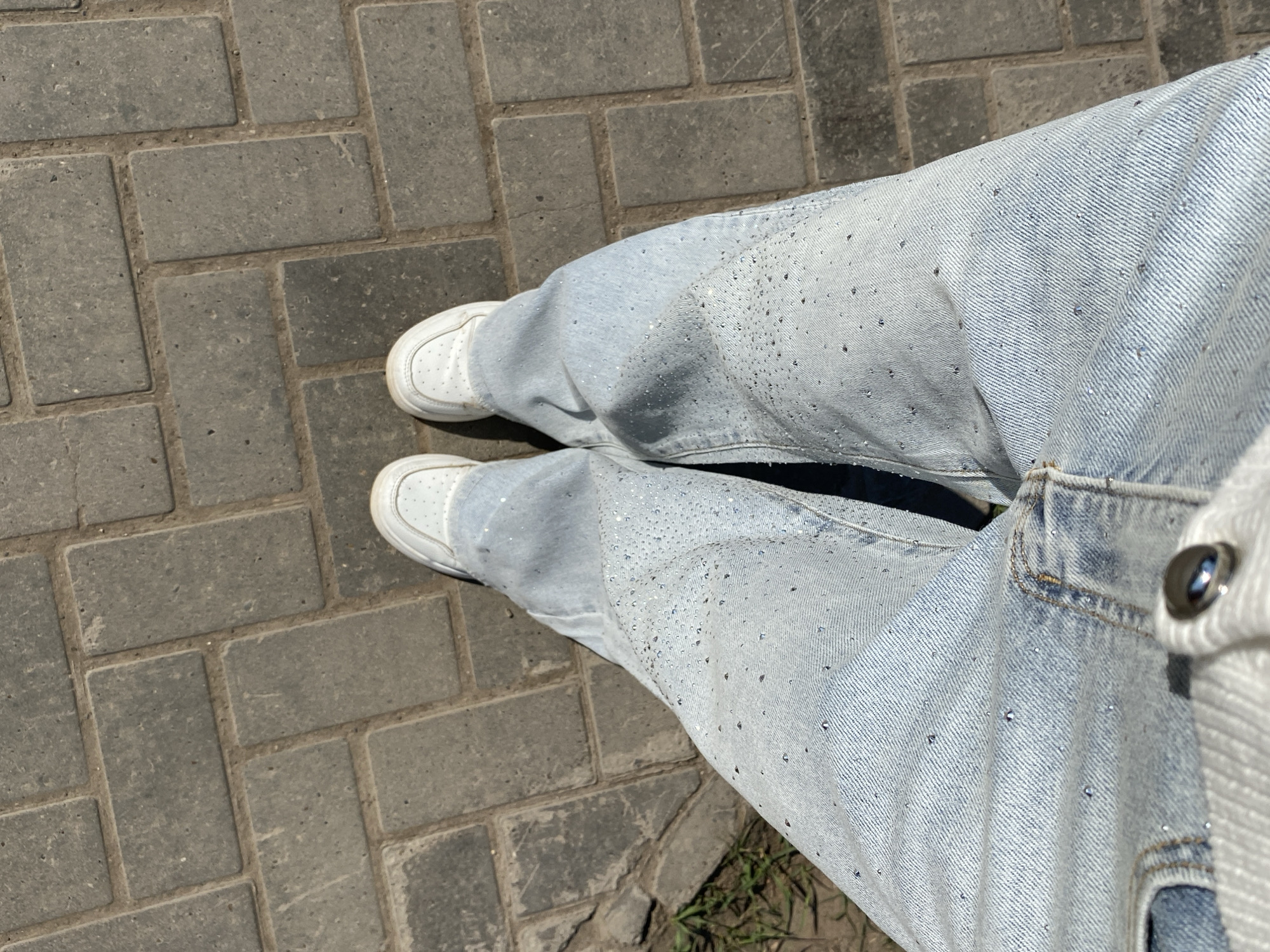 A first-person, eye-level shot shows a person standing on a grey brick sidewalk. The individual is wearing light-wash denim jeans adorned with scattered small, reflective embellishments. The jeans are paired with white sneakers. The location appears to be outdoors. The jeans have a button located in the lower right of the image. The sidewalk is formed from rectangular bricks arranged in a herringbone pattern. Patches of grass are visible at the bottom of the image. The scene is illuminated by natural sunlight.