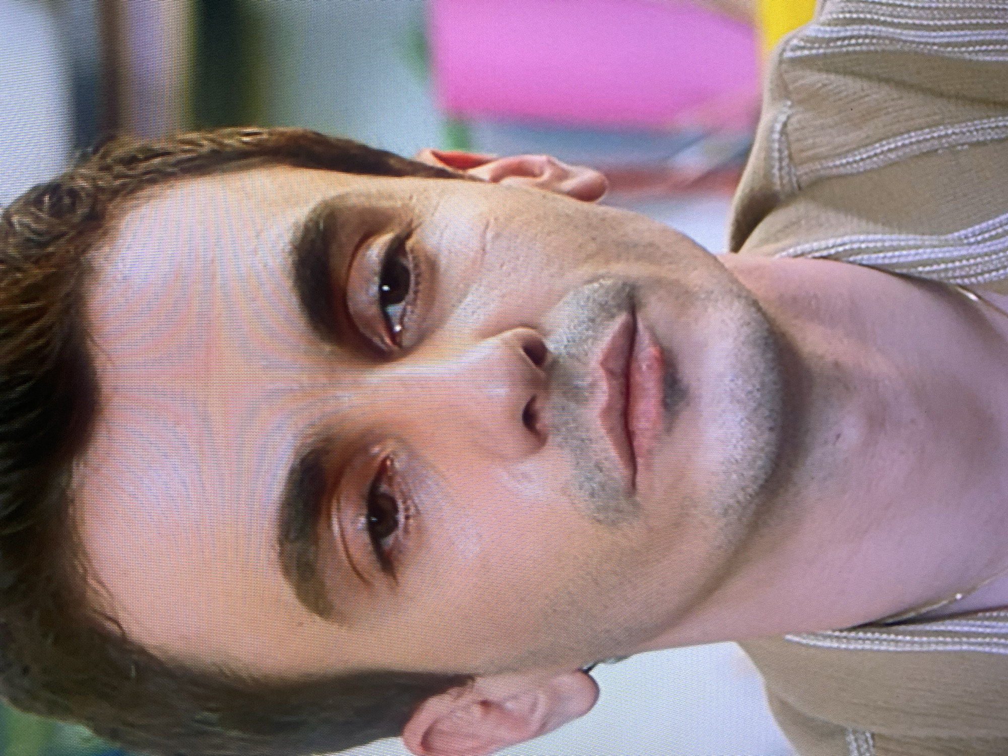 Close-up portrait of a man with dark brown hair, full eyebrows, brown eyes, and light skin. He has stubble on his face and appears to be wearing a beige and white striped shirt. The background is blurry with hints of pink and yellow. The location is not discernible from the image. The image provides no identifiable information regarding Ezerovo, Bulgaria.