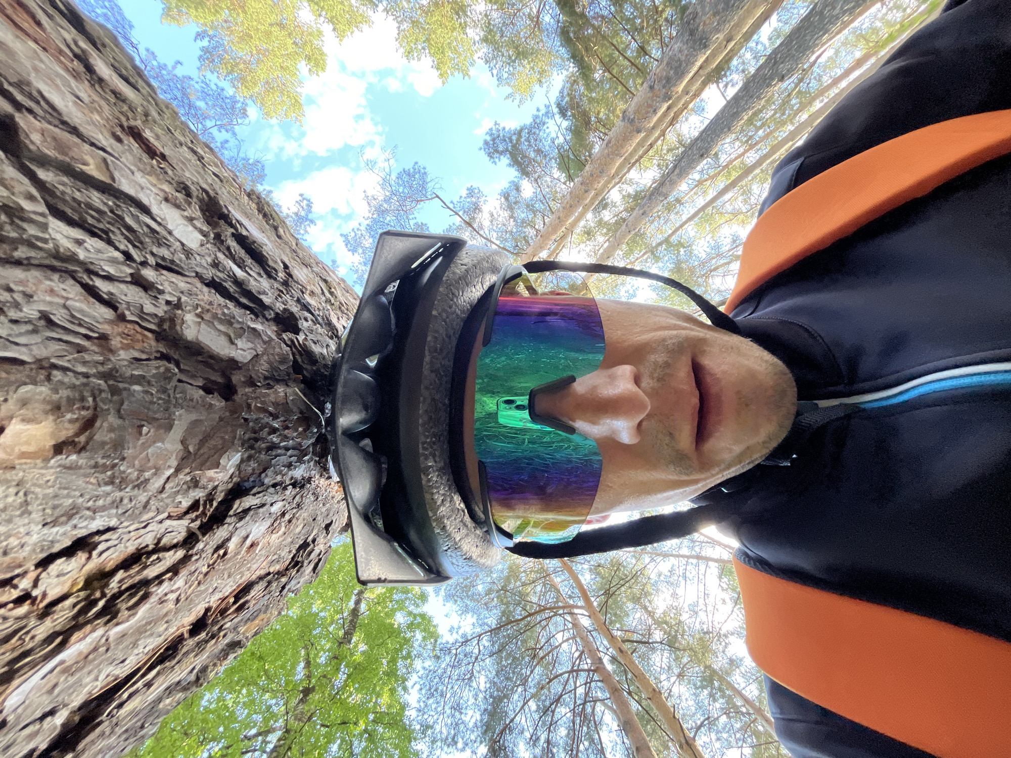 The image presents a low-angle, wide-angle perspective of a person in a forest, possibly a selfie taken from the ground looking upwards. The main subject is an individual, visible from the nose downwards, wearing a dark cycling helmet and large, iridescent wraparound sunglasses with a rainbow-like tint. A bright green smartphone with a camera module is clearly reflected in the right lens of the sunglasses.
The person wears a dark long-sleeved top, likely activewear such as a cycling jersey or jacket, with a prominent orange strap crossing their chest, suggestive of a backpack or hydration pack. A subtle blue stripe is visible on the collar area of the top. The expression on the visible part of the face is neutral, with the mouth slightly open.
The setting is a dense forest or wooded area, with tall trees dominating the background. Due to the extreme low angle, a large tree trunk with textured, rough bark appears to lie horizontally across the left side of the frame. Numerous other tree trunks and branches with green foliage fill the upper and right portions of the image, reaching towards a clear blue sky dotted with scattered white clouds. The trees appear to be conifers, possibly pines.
The activity taking place is implied to be an outdoor pursuit like cycling or hiking, given the helmet, sports sunglasses, and activewear in a natural environment. The bright, clear sky indicates good weather during the daytime. No visible text is clearly discernible on any of the subjects or objects within the frame. The location is in a forested area, consistent with the provided context of Chaikovsky, Russia.
