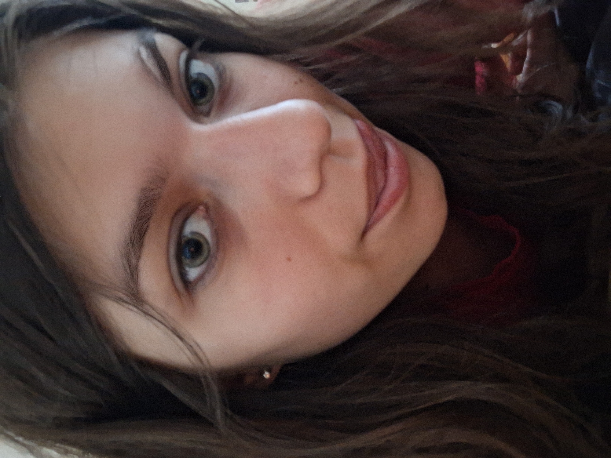 The image shows a young woman's face close-up. She has light skin, brown hair, and green eyes. A small mole is visible on her cheek. She wears a small gold earring. Her mouth is slightly open. The background is out of focus, showing brown hair and hints of red fabric. The image is angled, making it difficult to determine the precise orientation of the subject. There is no verifiable information that place the location in the city of Panagyurishte, Bulgaria.
