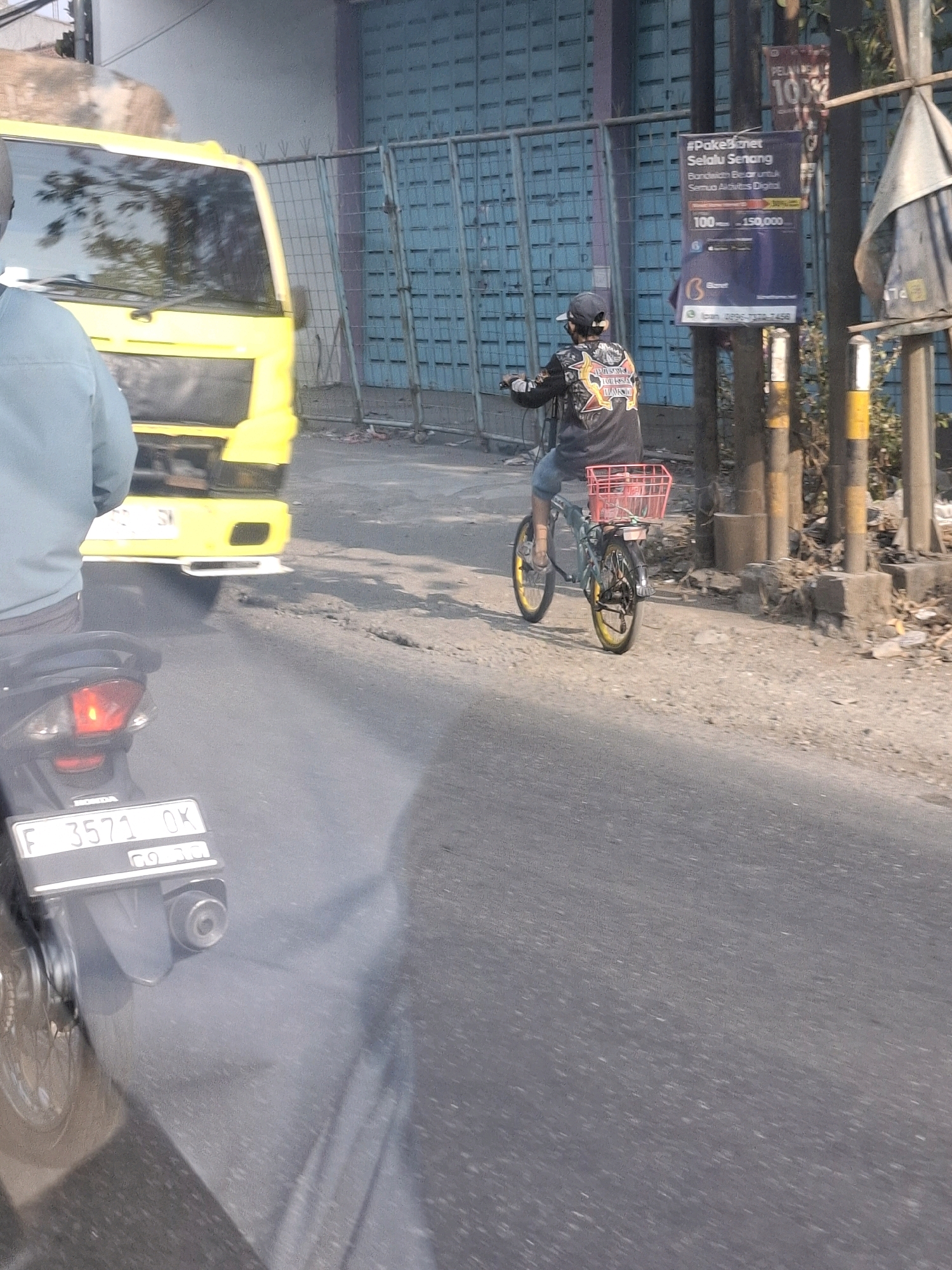 The image captures a street scene in Karangtengah, Indonesia, under bright daylight. In the foreground, a black motorcycle is partially visible, emitting a plume of exhaust smoke. Its license plate, reading "F 3571 OK", is clearly legible. To the left, a yellow truck is parked. Further down the road, a person wearing a black patterned jacket, a cap, and blue shorts is riding a bicycle equipped with a red basket. The bicycle has yellow rims. Behind the cyclist, a blue and white sign is affixed to a pole. The background shows a building with blue doors and some vegetation. The road surface is asphalt with visible cracks and debris.