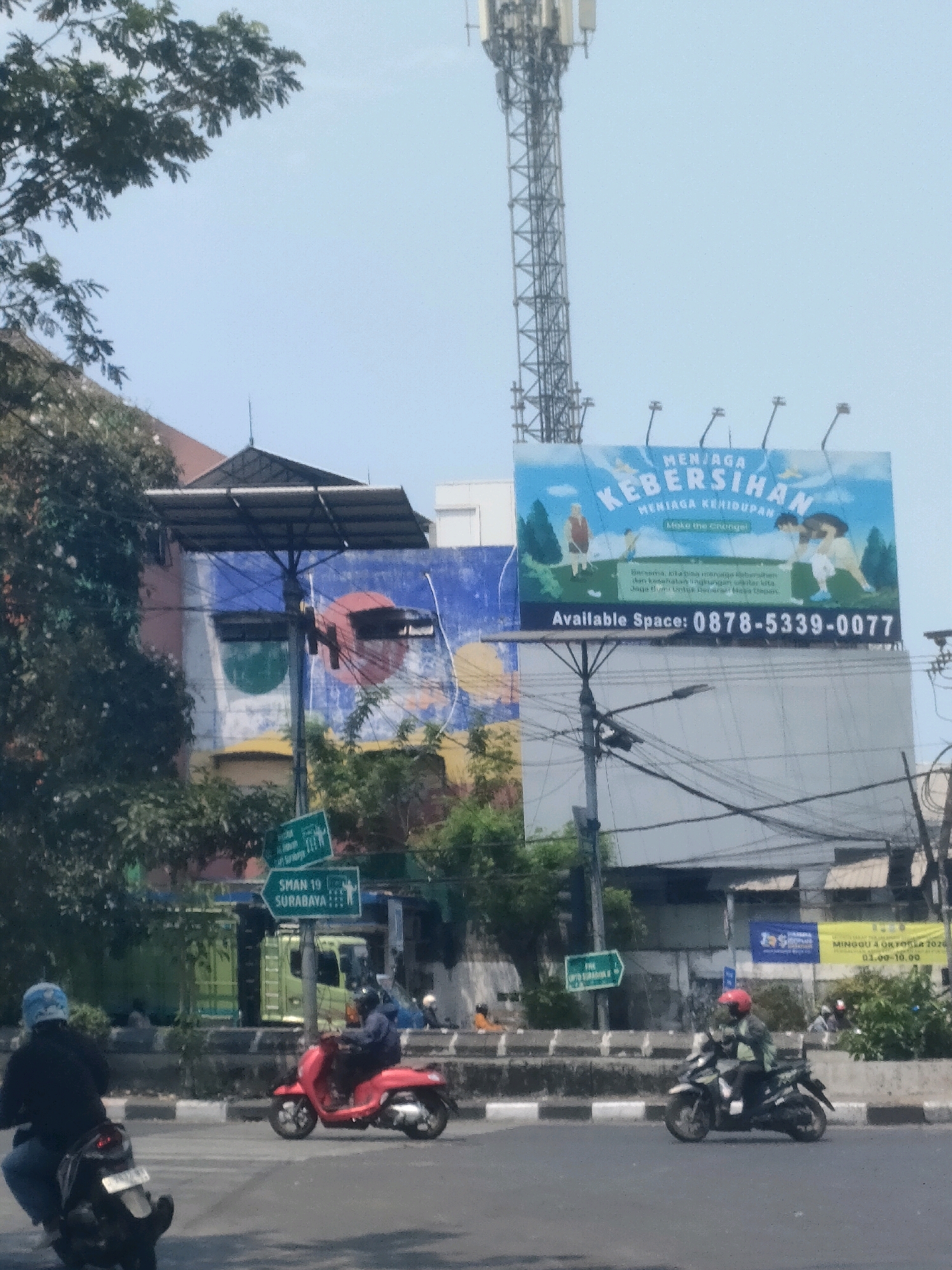 The image depicts a street scene in Surabaya, Indonesia, under a bright sky. A large billboard with the text "MENJAGA KEBERSIHAN MENJAGA KEHIDUPAN" and "Make the Change!" is prominently displayed, along with contact information "Available Space: 0878-5339-0077". Below the billboard, a green directional sign indicates "SMAN 19 SURABAYA" with a pedestrian icon. In the foreground, several motorcycles are visible. One motorcycle, a red scooter, is being ridden by a person wearing a dark jacket and a helmet. Another motorcycle, dark in color, is being ridden by a person in a black jacket and a blue helmet. A green truck is parked in the background, behind a concrete barrier. The environment includes buildings with colorful murals, trees, and a tall telecommunications tower in the distance. The overall scene captures a snapshot of urban life in Surabaya.