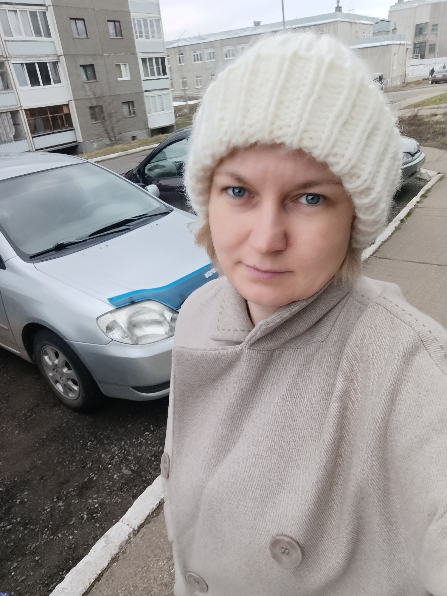 The image is a selfie of a light-skinned woman with blue eyes, wearing a thick white knit hat and a beige wool coat. She appears to be standing outdoors in a residential area, evidenced by the presence of a silver Toyota Corolla with a blue hood cover, a dark-colored sedan parked behind it, and a grey multi-story apartment building in the background. The ground is paved, and there's a white curb. The sky is overcast, suggesting a potentially cold or cloudy day. Based on geotags, the location is in Марковский, Russia.