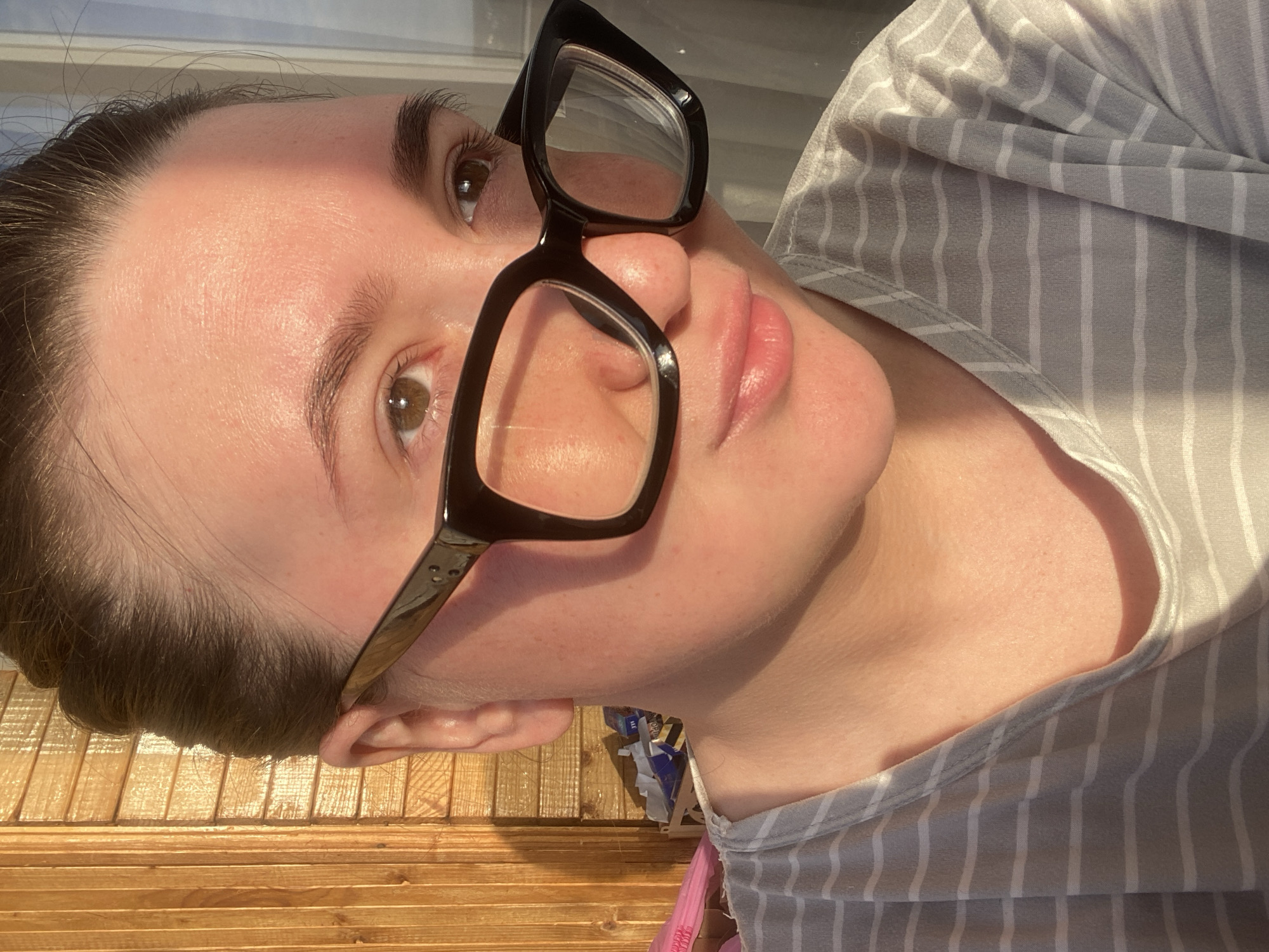 A close-up, rotated image depicts a person with fair skin, wearing black-framed glasses and a gray and white striped shirt. The person has brown hair pulled back and hazel eyes. The image is taken indoors. Due to the lack of contextual information within the image, specifying the location as the city of Chaikovsky, Russia, is not possible.