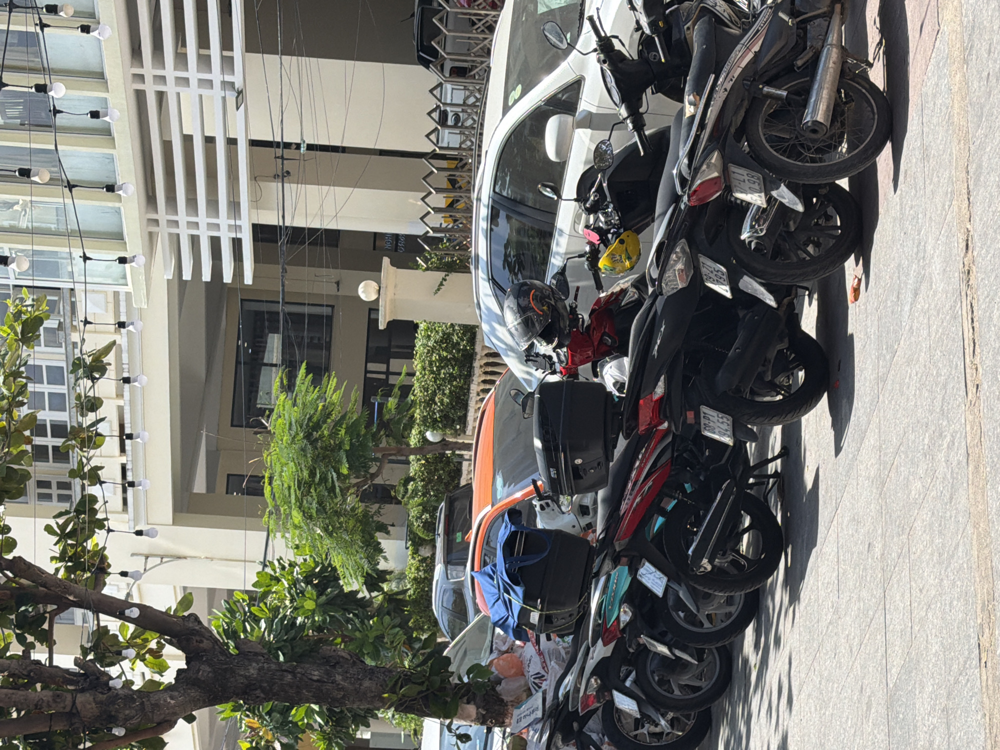The image shows a street scene in Bac Nha Trang Ward, Vietnam. A row of parked motorbikes lines the right side of the frame. A white car is parked among them. On the left is the facade of a building with decorative lighting. Trees are visible in the foreground and middle ground.
