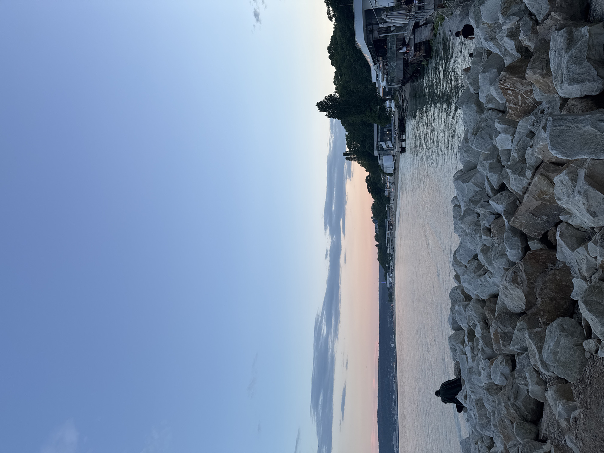 The image depicts a serene coastal scene at dusk, characterized by a clear sky transitioning from light blue at the zenith to soft pink and orange hues at the horizon, with sparse, elongated grey clouds in the mid-sky. The foreground is dominated by an extensive array of irregular, light-grey and white boulders, forming a rocky embankment or breakwater. In the mid-left, a solitary individual, dressed in dark clothing, is seated on these rocks, facing the tranquil, rippling expanse of the sea.
Across the water, a distant landmass features a dark, tree-covered shoreline, behind which a faint cityscape or town is visible. Along the immediate coastline on the right side of the image, several modern, white multi-story buildings, resembling restaurants or cafes, are visible. These structures feature outdoor wooden decks and patios extending over the water, some with white umbrellas providing shade. Lush green trees and vegetation densely cover the hillsides directly behind these buildings. Multiple small figures are discernible on and around these structures, indicating human presence. Another person in dark attire is visible standing near the water's edge on the far right, adjacent to the rocky foreground. The overall lighting suggests the soft ambient glow of early evening or sunset. The location is a coastal area featuring both natural rocky shorelines and developed commercial establishments by the water.