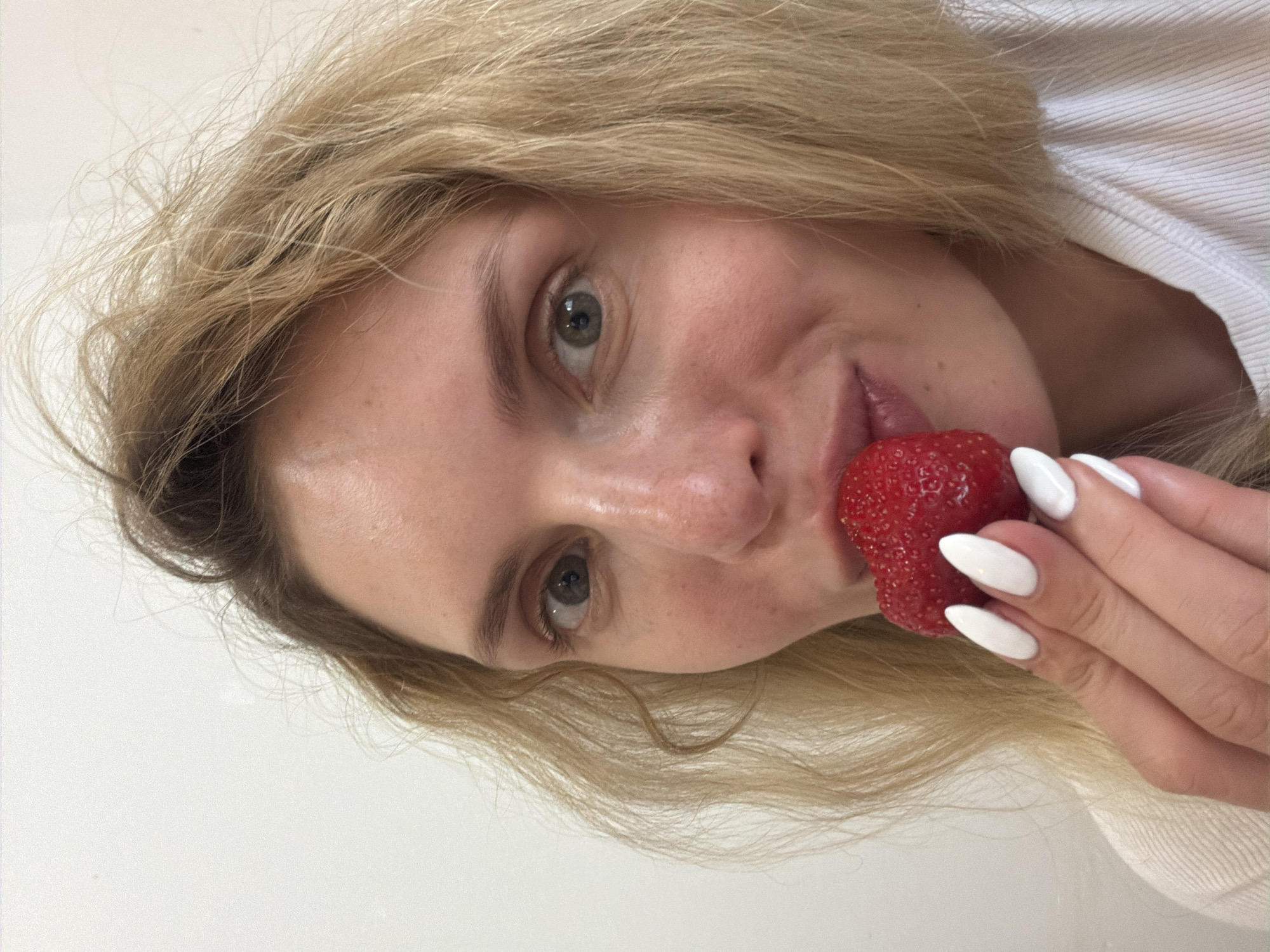A woman with fair skin, light blonde hair, and green eyes is holding a strawberry to her lips. She is wearing a white top. The background is a plain white surface. There is no indication of location within the city of Budva, Montenegro, or any identifiable features of the environment.