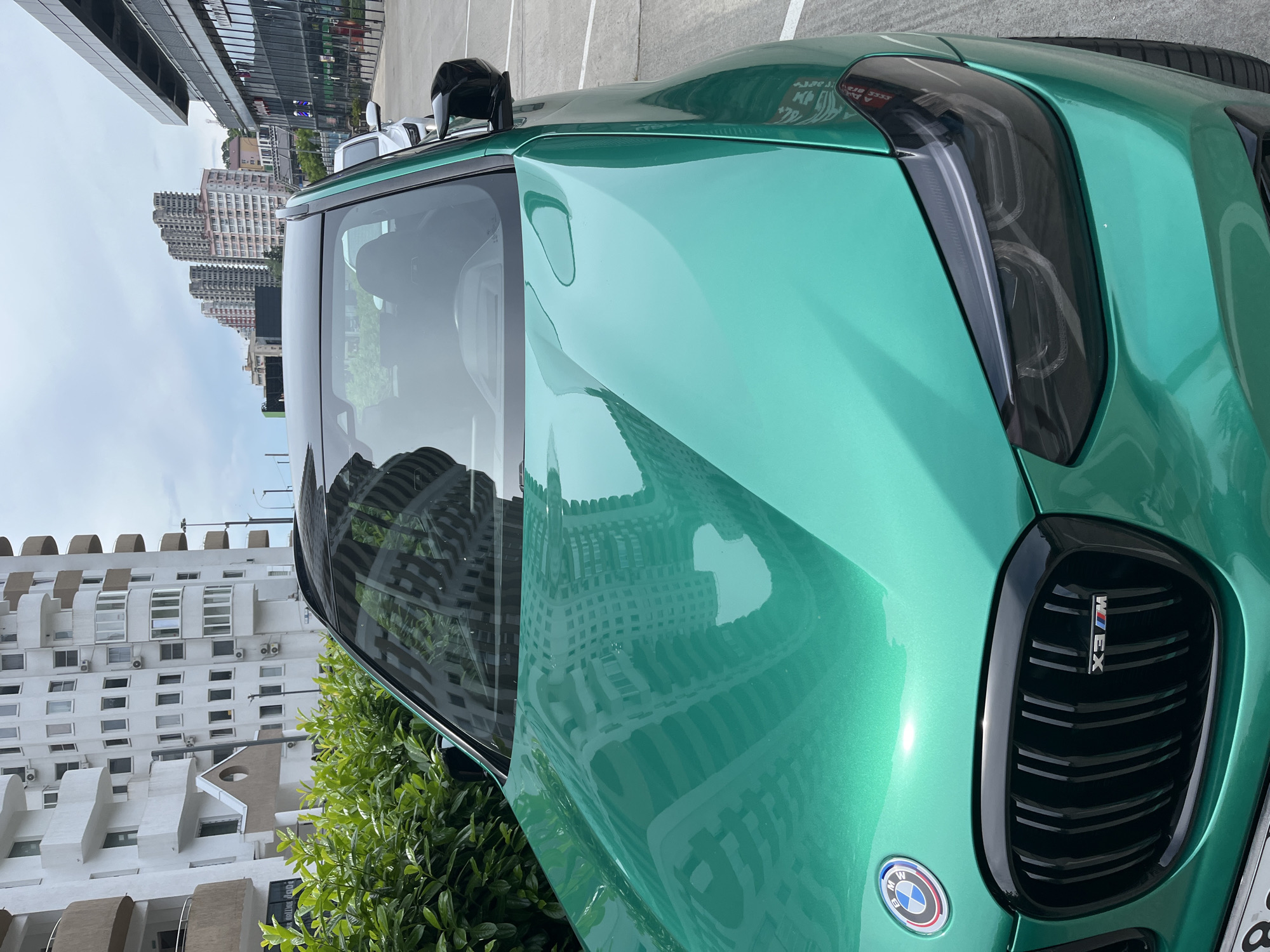 A bright green BMW X3 M is parked in an outdoor lot. The car's hood reflects a white building with balconies and large windows. In the background, a row of tall, modern apartment buildings is visible under a cloudy sky. The car's front grille and headlights are prominent. The location is the city of Sochi, Russia.