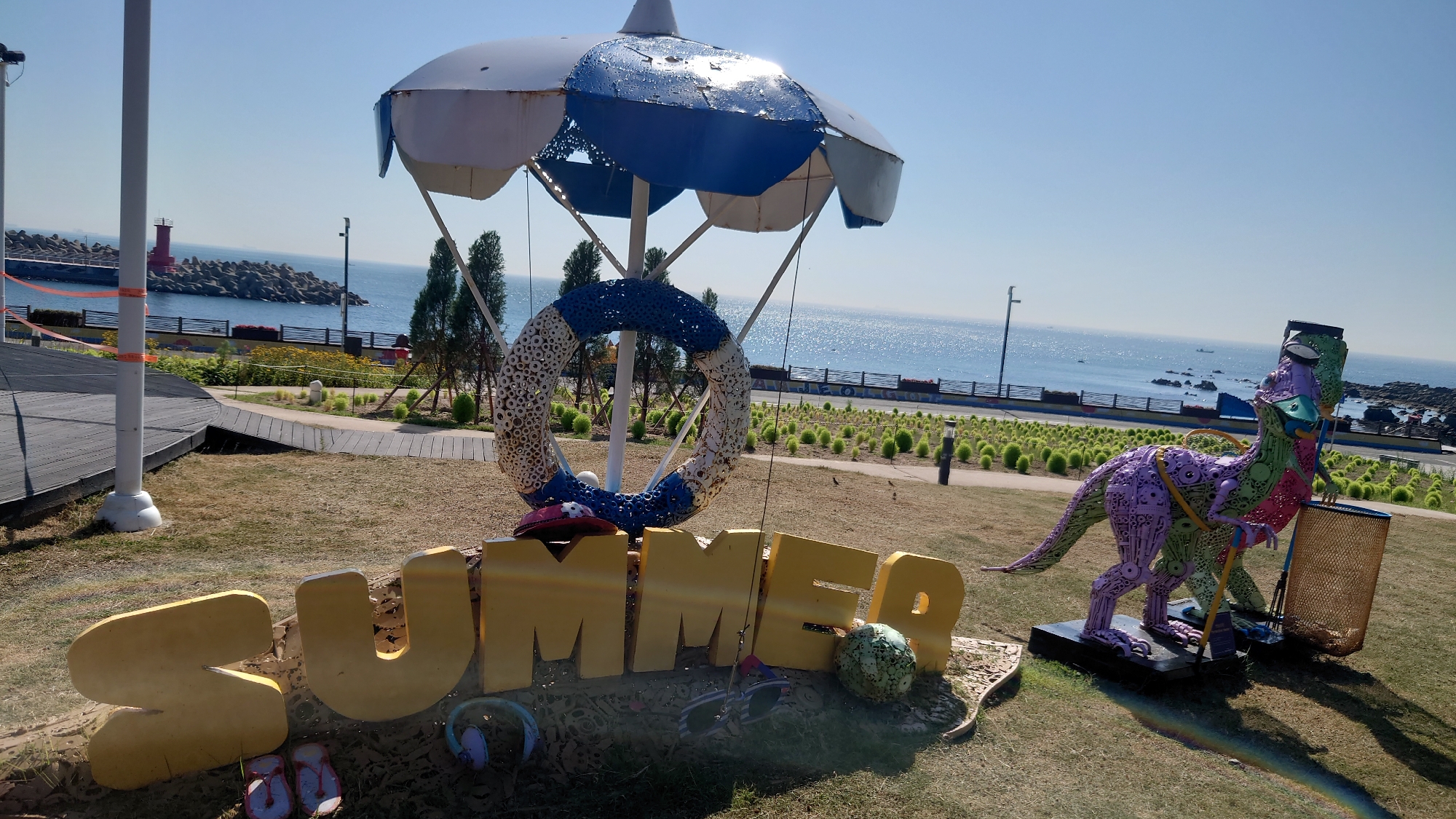 This image depicts a festive outdoor decoration near the coast, likely in Ulsan, South Korea, given the presence of a lighthouse and breakwater characteristic of the region. In the foreground, large, golden, three-dimensional letters spell out "SUMMER." Beneath the letters, a pair of pink flip-flops and a blue object, possibly headphones, are placed. To the right of the "SUMMER" sign, a colorful, metallic dinosaur sculpture stands, painted in shades of purple and green, with a metal net bin behind it. The dinosaur is leaning on a golf club and appears to be wearing sunglasses. Behind the "SUMMER" sign, a large, weathered umbrella-like structure is prominently featured, adorned with a circular rope decoration, which resembles a lifebuoy. The background reveals a clear blue sky and the expansive ocean, with a breakwater and a red lighthouse visible on the left. The area is landscaped with dry grass and rows of rounded bushes, suggesting a park or a public recreational space along the coastline. The overall scene conveys a summery and playful atmosphere.