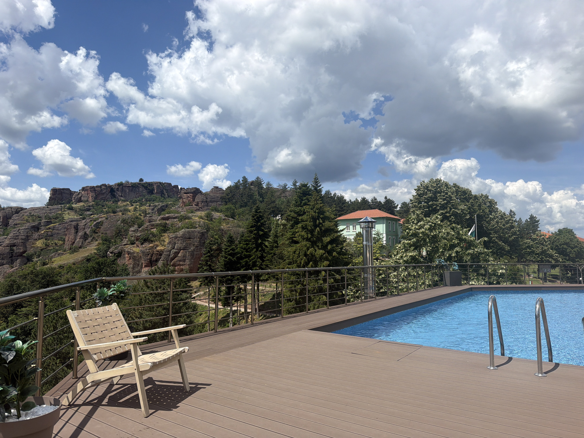 The image depicts a sunny day with scattered clouds over a natural landscape in Belogradchik, Bulgaria. In the foreground, a wooden deck with a modern swimming pool is visible. A light-colored lounge chair sits on the deck, angled towards a panoramic view. The deck surrounds the blue water of the pool, which features metal ladder handrails. Beyond the pool area, a railing separates the deck from a natural embankment.
The embankment is covered in dense green trees and rugged, reddish-brown rock formations characteristic of the Belogradchik Rocks. A light green building with a red roof is situated amidst the trees on the hillside, suggesting a residential or hospitality establishment. The sky is a vibrant blue, dotted with fluffy white clouds, some of which are large and dramatic. The overall scene conveys a sense of natural beauty and a relaxing outdoor setting.