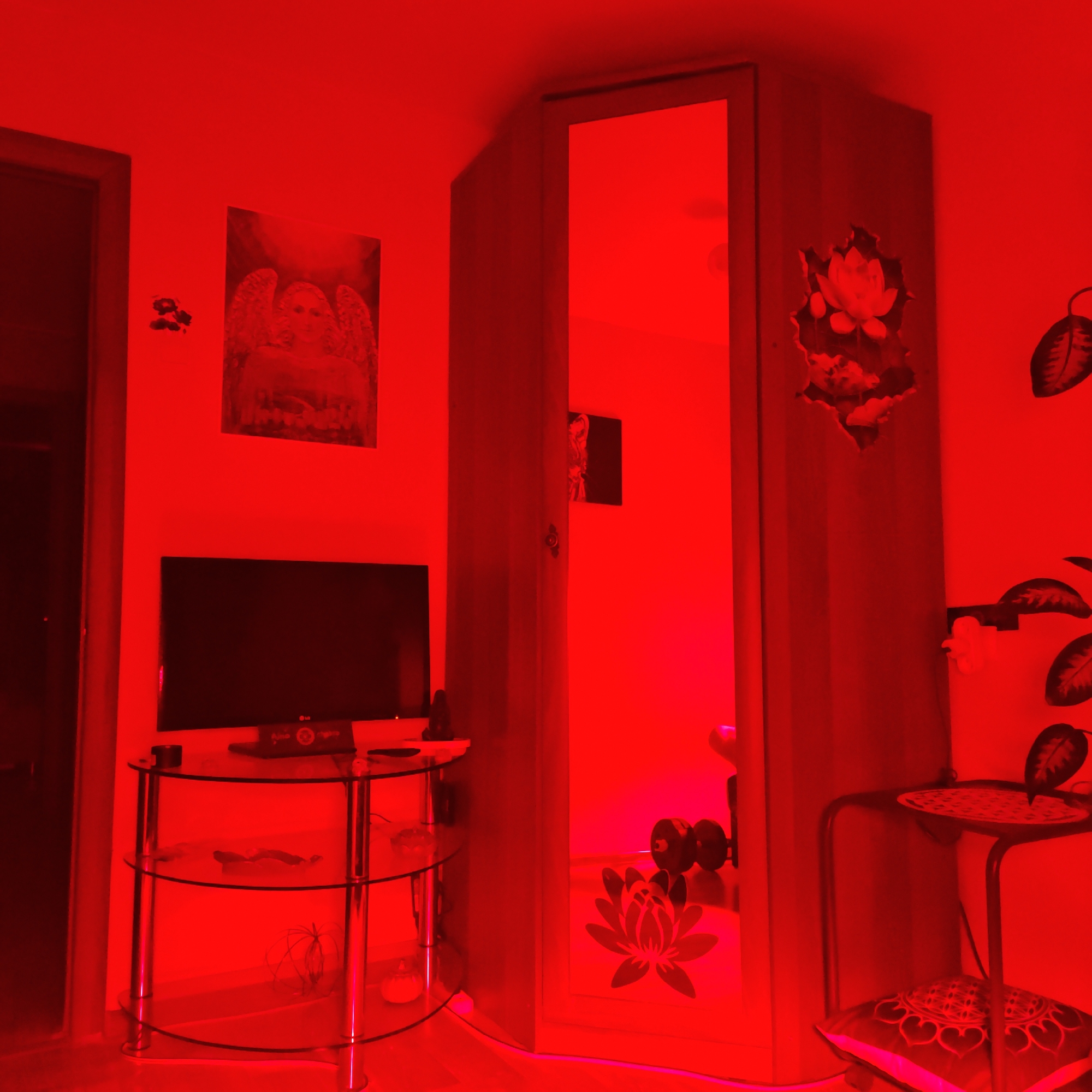 The image shows an interior space, likely a room in a building. The lighting is dominated by a strong red hue.
Furniture and objects present include:
* A television set.
* A glass-shelved table or stand beneath the television.
* A wardrobe or cabinet with a mirror panel. The mirror reflects a set of dumbbells inside.
* A small side table with a patterned cushion placed underneath.
* Wall art, including what appears to be a religious depiction and other decorative elements.
The location is identified as Rostov, Russia. This is only a place of reference and the scene shown could be anywhere. No people are visible. The overall impression is a domestic setting, though the strong red lighting obscures specific details of the objects and the general environment.