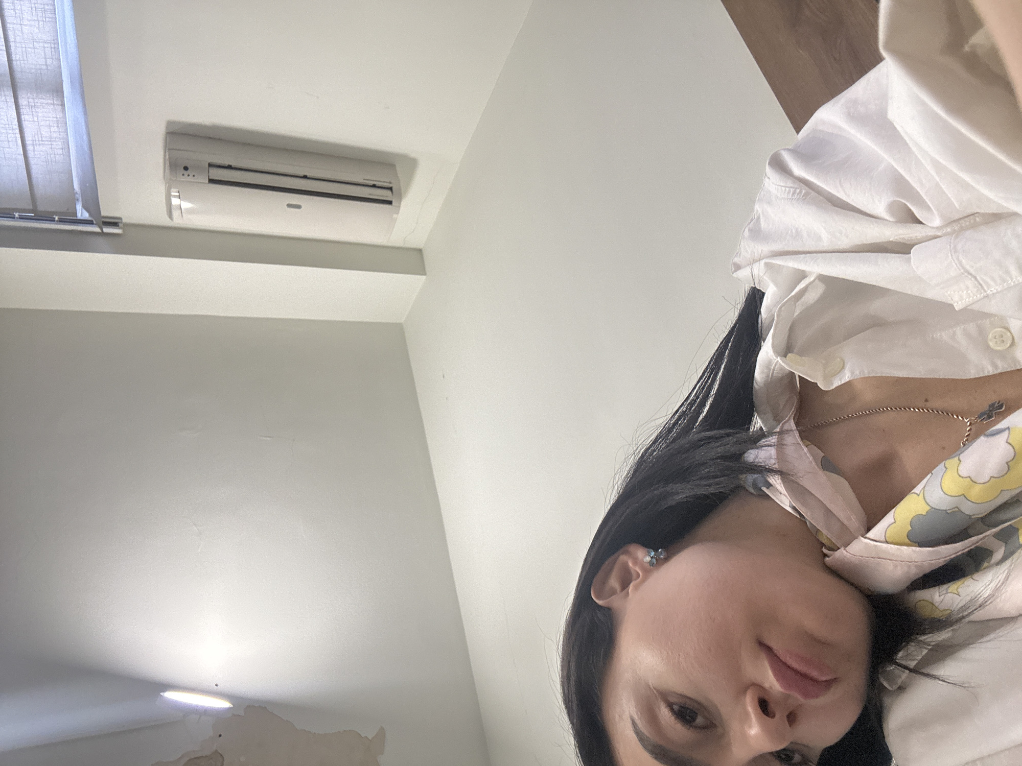 A woman with dark hair is looking down at the camera. She is wearing a white collared shirt, with a yellow and grey pattern visible on a garment underneath. She has a necklace with a cross pendant. Behind her is a white wall with an air conditioning unit. To the left, there is a window with blinds. A light fixture is visible on the wall in the lower left of the frame. The location is likely indoors, potentially in Tbilisi, Georgia.