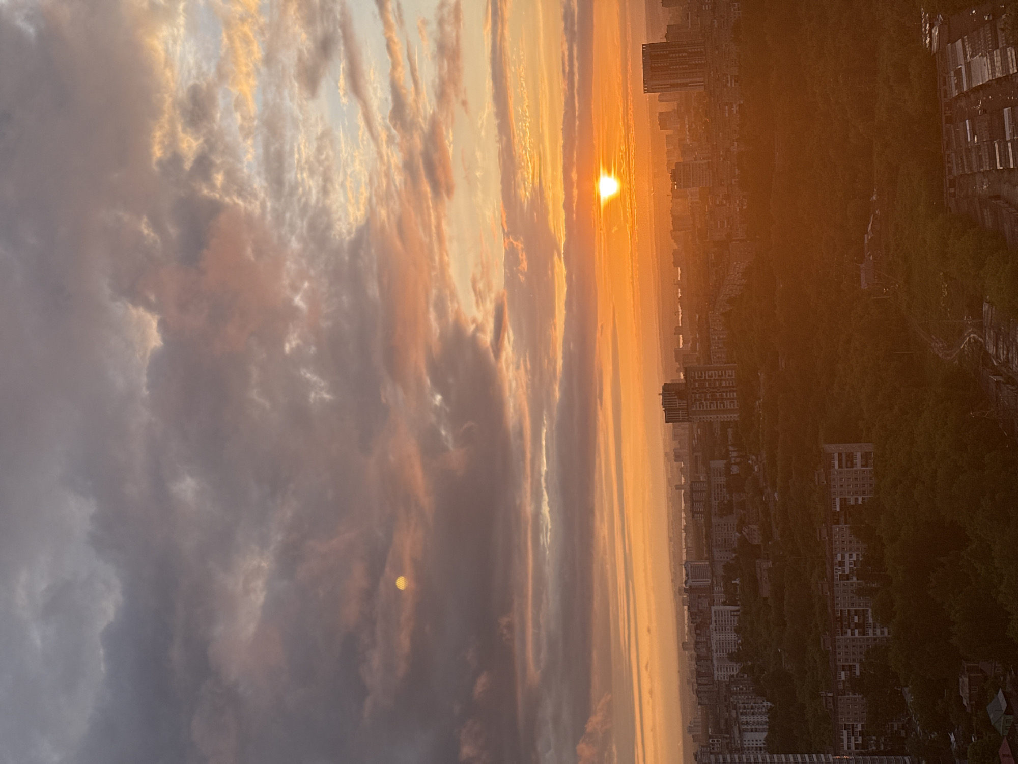 An elevated wide-angle photograph depicts a city at sunset or sunrise. The sky occupies the upper two-thirds, filled with extensive stratocumulus clouds exhibiting hues of intense orange, yellow, pink, and grey, illuminated from below. A brilliant, partially obscured sun, appearing as a distinct luminous disc, is located near the right-center horizon, casting a strong golden glare across the lower sky and atmosphere. In the upper-left quadrant, a smaller, circular, yellowish-orange object with a distinct grid-like pattern is visible, consistent with a lens flare.
The urban landscape below comprises a dense mix of high-rise and multi-story residential buildings, many exhibiting post-Soviet architectural styles, interspersed with extensive areas of dark green foliage indicative of parks or wooded zones. A hazy body of water is discernible on the distant horizon beyond the city. No individuals or specific activities are visible within the frame. The image lacks specific geographic identifiers to confirm its location as Гамово, Russia.
